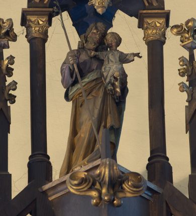 Zdjęcie nr 1: Rzeźba wolnostojąca, przedstawiająca św. Józefa z Dzieciątkiem. Święty jest ukazany w postawie stojącej, w delikatnym kontrapoście z Dzieciątkiem Jezus na lewym ręku oraz gałązką lilii w prawej, z głową przechyloną na swoją, lewą stronę. Ma podłużną twarz, z długim i szerokim nosem, z wzrokiem skierowanym w dal, okoloną krótką brodą i długimi, bujnymi włosami. Ubrany jest w fioletową suknię z długimi rękawami oraz złocony płaszcz z zieloną podszewką. Dzieciątko Jezus siedzi na przedramieniu św. Józefa, lewą nóżkę wysuwa do przodu, a dłonie rozpościera szeroko na boki. Ma szeroką twarz o dziecięcych rysach, okoloną krótkimi i jasnymi włosami. Ubrane jest w długą i białą sukienkę z długimi rękawami. 