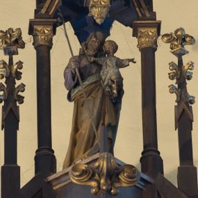 Zdjęcie nr 1: Rzeźba wolnostojąca, przedstawiająca św. Józefa z Dzieciątkiem. Święty jest ukazany w postawie stojącej, w delikatnym kontrapoście z Dzieciątkiem Jezus na lewym ręku oraz gałązką lilii w prawej, z głową przechyloną na swoją, lewą stronę. Ma podłużną twarz, z długim i szerokim nosem, z wzrokiem skierowanym w dal, okoloną krótką brodą i długimi, bujnymi włosami. Ubrany jest w fioletową suknię z długimi rękawami oraz złocony płaszcz z zieloną podszewką. Dzieciątko Jezus siedzi na przedramieniu św. Józefa, lewą nóżkę wysuwa do przodu, a dłonie rozpościera szeroko na boki. Ma szeroką twarz o dziecięcych rysach, okoloną krótkimi i jasnymi włosami. Ubrane jest w długą i białą sukienkę z długimi rękawami. 