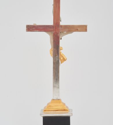 Zdjęcie nr 3: Podstawa w formie prostego prostopadłościanu na niskim cokoliku, zwieńczona gzymsem, wyżej przechodząca w zwężający się, profilowany cokół na planie prostokąta, na którym osadzony jest krucyfiks. Ramiona krzyża ujęte przy krawędzi bordową opaską, wewnątrz złocone i karbowane. Figura Chrystusa ścięta z tyłu, przybita do krzyża trzema gwoździami. Ciało w delikatnym zwisie o szeroko rozłożonych ramionach, głowa opada na prawe ramię, nogi są ugięte w kolanach, kolana przylegają do siebie, a stopy są przebite w układzie prawa na lewą. Twarz szczupła o ostrym profilu nosa, okolona krótką brodą. Włosy długie, silnie skręcone w pukle i opadające na tył głowy i prawe ramię. Ciało szczupłe o delikatnie podkreślonej muskulaturze oraz uwypuklonej formie klatki piersiowej, szczególnie linii żeber z zaznaczonymi śladami męki. Na głowie korona cierniowa. Przez biodra przewiązane jest złocone perizonium w formie krótkiej skręconej tkaniny, zawieszonej na sznurze, odsłaniającej prawe biodro, ze zwisem na prawym boku. Na zakończeniu pionowej belki titulus w formie zwoju z napisem „IN / RI”. Polichromia w odsłoniętych partiach ciała Chrystusa delikatnie sina, perizonium, krzyż i detale złocone oraz srebrzone, postument polichromowany na czarno. 
