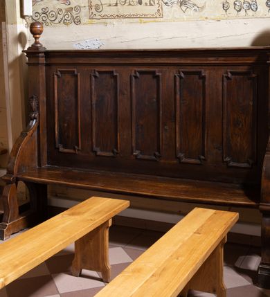 Zdjęcie nr 2: Drewniane ławki kolatorskie ustawione po obu stronach prezbiterium. Ławki ustawione na dwóch nóżkach, siedzisko proste w postaci prostej ławy. Zaplecek flankowany filarkami podtrzymującymi wyłamane, niepełne belkowanie, zamknięte gzymsem. Na osi filarków ustawione wazony. Zaplecek zdobiony pięcioma płycinami w kształcie stojących prostokątów o uszatych narożach. Ścianki boczne z przodu o wklęsło-wypukłym brzegu, sięgające do wysokości połowy zaplecka, na zakończeniu zdobione liśćmi stylizowanego akantu. Struktura pokryta bejcą.
