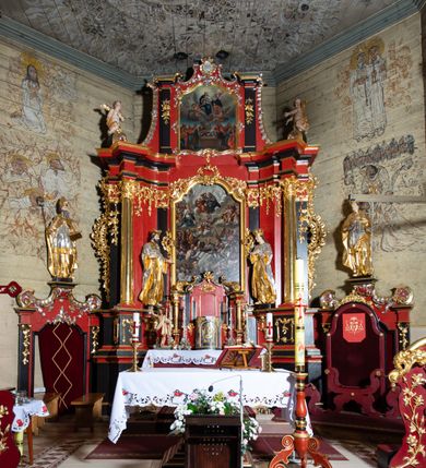 Zdjęcie nr 6: Nastawa architektoniczna, dwukondygnacyjna, jednoosiowa z bramkami, na dwustrefowym cokole. Ołtarz kamienny, prostopadłościenny, w drewnianej obudowie, mensa drewniana z marmurowym portatylem. Antepedium w formie leżącego prostokąta ujętego profilowaną ramą; pole wyłożone tkaniną o kwiatowym wzorze z haftowanym medalionem w glorii z monogramem IHS; ujęty gałązkami lilii. Tabernakulum w formie drewnianej skrzynki z wypukłym frontem, obite srebrzoną blachą. Drzwiczki w kształcie stojącego prostokąta zamkniętego łukiem półkolistym, obwiedzione ramą. W polu aplikacja kielicha z hostią w glorii, ujętego kłosami zbóż; w przyłuczach ornament roślinny. Tabernakulum w architektonicznej obudowie tworzącej tron wystawienia; ujęte parą postumentów dekorowanych rocaille'owymi płycinami oraz parą filarów, przed którymi stoi para kolumn na postumentach utworzonych z ornamentalnych konsol. Podpory dźwigają belkowanie, wyłamane pośrodku, przerwane nad tabernakulum. Za tabernakulum niewielka ścianka zamknięta szczytem utworzonym z dwóch spływów wolutowych. Ze szczytu na belkowanie spływają ornamentalne woluty. Tron ujęty rocaille'owymi uszami. 
Pierwsza kondygnacja ujęta grupą zwielokrotnionych pilastrów, przed którymi stoją ustawione pod kątem cokoły z kolumnami o stylizowanych kapitelach kompozytowych, dźwigających pełne belkowanie, przerwane nad polem środkowym. Na tle pilastrów figury św. Jana Kantego po lewej stronie i św. Kazimierza po prawej. W polu środkowym obraz „Wszyscy Święci” ujęty profilowaną ramą w formie stojącego prostokąta, zamkniętego łukiem wklęsło-wypukłym z uskokami. Na zasuwie obraz przedstawiający Ukrzyżowanie Chrystusa. Druga kondygnacja w formie zbliżonej do kwadratu, flankowana przez zdwojone pilastry i spływy wolutowe, zamknięte szczytem z rocaille'owym kartuszem z napisem „OPŁUKALI / SWE SZATY / W KRWI / BARANKA / AP 7,14”. W polu obraz „Wniebowzięcie Najświętszej Marii Panny” ujęty profilowaną ramą w formie zbliżonej do kwadratu od dołu i od góry o wklęsło-wypukłym zarysie. Na zakończeniu spływów wolutowych usadzone dwie figury aniołów. Bramki w formie stojącego prostokąta ujętego parą pilastrów, dźwigających spływy wolutowe, podtrzymujące postumenty dla figur św. Stanisława po lewej stronie i św. Wojciecha po prawej. Przejścia w formie stojących prostokątów, zamkniętych stylizowanymi łukami segmentowymi nadwieszonymi. Całość dekorowana ornamentem rocaille'owym oraz różanymi zwisami w partii trzonów podpór, pierwsza kondygnacja flankowana rocaille'owymi, ażurowymi uszami. Struktura polichromowana w kolorze czerwonym, z malowanymi na czarno trzonami pilastrów i profilami, złoconymi kolumnami, profilami i ornamentyką oraz srebrzonymi detalami. Według dokumentacji konserwatorskiej na odwrociach baz kolumn znajdują się napisy „Leonora Olajassy żona organisty. 1887” oraz „Leonora Olajassy - organiścina. 1887”.