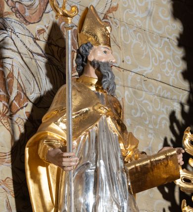 Zdjęcie nr 1: Rzeźba ścięta z tyłu, drążona, przedstawiająca św. Wojciecha. Święty ustawiony frontalnie w kontrapoście, z lewą nogą ugiętą w kolanie. Głowę zwraca w swoją lewą stronę, w prawej dłoni trzyma pastorał, a w lewej księgę. Twarz szczupła o silnie uwypuklonych kościach policzkowych z dużym nosem, migdałowymi oczami i otwartymi ustami; okolona długą i siwą brodą, opadającą falowanymi pasmami. Włosy krótkie i kręcone. Ubrany jest w długą albę obficie drapowaną u spodu, rokietę z dekoracyjnie obszytym brzegiem i kapę zapiętą na piersi. Na stopach ma założone złocone buty, a na głowie infułę. Cokół niski i prostopadłościenny, pomalowany na ciemnobrązowo. Polichromia naturalistyczna w odsłoniętych partiach ciała, strój i atrybuty złocone i srebrzone.