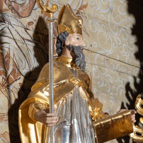 Zdjęcie nr 1: Rzeźba ścięta z tyłu, drążona, przedstawiająca św. Wojciecha. Święty ustawiony frontalnie w kontrapoście, z lewą nogą ugiętą w kolanie. Głowę zwraca w swoją lewą stronę, w prawej dłoni trzyma pastorał, a w lewej księgę. Twarz szczupła o silnie uwypuklonych kościach policzkowych z dużym nosem, migdałowymi oczami i otwartymi ustami; okolona długą i siwą brodą, opadającą falowanymi pasmami. Włosy krótkie i kręcone. Ubrany jest w długą albę obficie drapowaną u spodu, rokietę z dekoracyjnie obszytym brzegiem i kapę zapiętą na piersi. Na stopach ma założone złocone buty, a na głowie infułę. Cokół niski i prostopadłościenny, pomalowany na ciemnobrązowo. Polichromia naturalistyczna w odsłoniętych partiach ciała, strój i atrybuty złocone i srebrzone.