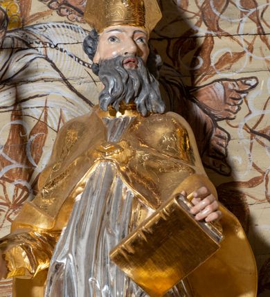 Zdjęcie nr 4: Rzeźba ścięta z tyłu, drążona, przedstawiająca św. Wojciecha. Święty ustawiony frontalnie w kontrapoście, z lewą nogą ugiętą w kolanie. Głowę zwraca w swoją lewą stronę, w prawej dłoni trzyma pastorał, a w lewej księgę. Twarz szczupła o silnie uwypuklonych kościach policzkowych z dużym nosem, migdałowymi oczami i otwartymi ustami; okolona długą i siwą brodą, opadającą falowanymi pasmami. Włosy krótkie i kręcone. Ubrany jest w długą albę obficie drapowaną u spodu, rokietę z dekoracyjnie obszytym brzegiem i kapę zapiętą na piersi. Na stopach ma założone złocone buty, a na głowie infułę. Cokół niski i prostopadłościenny, pomalowany na ciemnobrązowo. Polichromia naturalistyczna w odsłoniętych partiach ciała, strój i atrybuty złocone i srebrzone.