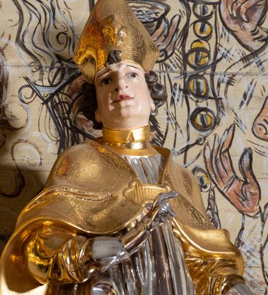 Zdjęcie nr 4: Rzeźba ścięta z tyłu, drążona, przedstawiająca św. Stanisława Biskupa. Święty ustawiony frontalnie, w delikatnym kontrapoście, w lewej ręce trzyma pastorał, prawą ugiętą w łokciu czyni gest błogosławieństwa. Twarz szeroka o silnie zaznaczonym podbródku, dużym nosie, ściągniętych brwiach, dużych oczach patrzących na wprost oraz niewielkich, rozchylonych ustach. Włosy krótkie, ciemnobrązowe i silnie skręcone po bokach głowy. Święty ubrany jest w albę, obficie drapowaną u spodu; rokietę z dekoracyjnym obszyciem u brzegu; kapę zapiętą na piersi; złocone buty, rękawice i infułę na głowie. Polichromia naturalistyczna w odsłoniętych partiach ciała, szaty i pastorał złocone i srebrzone.