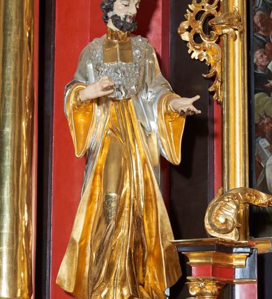 Zdjęcie nr 2: Rzeźba św. Jana Kantego, ścięta z tyłu, drążona. Święty ustawiony frontalnie w delikatnym kontrapoście z prawą nogą ugiętą w kolanie, ręce zgięte w łokciach, dłonie ułożone w teatralnych gestach. Twarz trójkątna, o uwypuklonych kościach policzkowych, dużym i bulwiastym nosie, wąskich oczach oraz niewielkich ustach; okolona krótką, gęstą i kręconą brodą. Włosy krótkie, skręcone w pukle po bokach głowy. Święty ubrany jest w długą złoconą suknię, przylegającą do ciała; togę rektorską o szerokich rękawach, rozchylającą się na boki, ze srebrzoną załogą (pelerynką) i złoconą befką; na stopach pełne buty. Polichromia naturalistyczna w odsłoniętych partiach ciała, szaty złocone i srebrzone.
