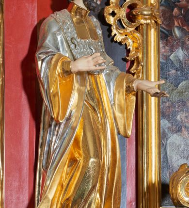 Zdjęcie nr 3: Rzeźba św. Jana Kantego, ścięta z tyłu, drążona. Święty ustawiony frontalnie w delikatnym kontrapoście z prawą nogą ugiętą w kolanie, ręce zgięte w łokciach, dłonie ułożone w teatralnych gestach. Twarz trójkątna, o uwypuklonych kościach policzkowych, dużym i bulwiastym nosie, wąskich oczach oraz niewielkich ustach; okolona krótką, gęstą i kręconą brodą. Włosy krótkie, skręcone w pukle po bokach głowy. Święty ubrany jest w długą złoconą suknię, przylegającą do ciała; togę rektorską o szerokich rękawach, rozchylającą się na boki, ze srebrzoną załogą (pelerynką) i złoconą befką; na stopach pełne buty. Polichromia naturalistyczna w odsłoniętych partiach ciała, szaty złocone i srebrzone.
