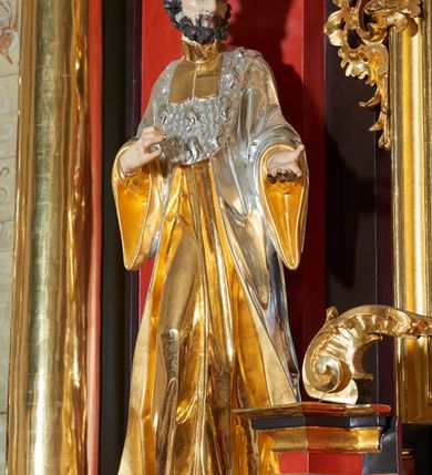 Zdjęcie nr 1: Rzeźba św. Jana Kantego, ścięta z tyłu, drążona. Święty ustawiony frontalnie w delikatnym kontrapoście z prawą nogą ugiętą w kolanie, ręce zgięte w łokciach, dłonie ułożone w teatralnych gestach. Twarz trójkątna, o uwypuklonych kościach policzkowych, dużym i bulwiastym nosie, wąskich oczach oraz niewielkich ustach; okolona krótką, gęstą i kręconą brodą. Włosy krótkie, skręcone w pukle po bokach głowy. Święty ubrany jest w długą złoconą suknię, przylegającą do ciała; togę rektorską o szerokich rękawach, rozchylającą się na boki, ze srebrzoną załogą (pelerynką) i złoconą befką; na stopach pełne buty. Polichromia naturalistyczna w odsłoniętych partiach ciała, szaty złocone i srebrzone.
