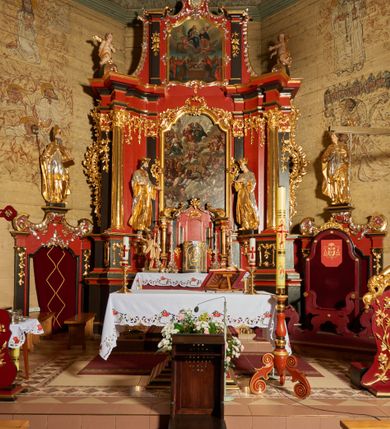 Zdjęcie nr 5: Rzeźba św. Jana Kantego, ścięta z tyłu, drążona. Święty ustawiony frontalnie w delikatnym kontrapoście z prawą nogą ugiętą w kolanie, ręce zgięte w łokciach, dłonie ułożone w teatralnych gestach. Twarz trójkątna, o uwypuklonych kościach policzkowych, dużym i bulwiastym nosie, wąskich oczach oraz niewielkich ustach; okolona krótką, gęstą i kręconą brodą. Włosy krótkie, skręcone w pukle po bokach głowy. Święty ubrany jest w długą złoconą suknię, przylegającą do ciała; togę rektorską o szerokich rękawach, rozchylającą się na boki, ze srebrzoną załogą (pelerynką) i złoconą befką; na stopach pełne buty. Polichromia naturalistyczna w odsłoniętych partiach ciała, szaty złocone i srebrzone.
