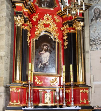 Zdjęcie nr 8: Nastawa architektoniczna, jednokondygnacyjna, jednoosiowa ze zwieńczeniem. Ołtarz drewniany, prostopadłościenny. Antepedium w kształcie leżącego prostokąta, ujęte profilowaną, polichromowaną, złoconą i srebrzoną ramą z ornamentyką roślinną. Pole wypełnione czerwoną tkaniną z haftem z gorejącym sercem oplecionym koroną cierniową pośrodku, flankowanym parą przeciwstawnie ustawionych gałązek białej lilii. Tabernakulum w kształcie prostopadłościennej skrzynki na niskim cokole, z lekko wypukłą ścianką przednią, flankowaną parą pilastrów z przylegającymi do nich półkolumienkami wspierającymi wyłamujący się ponad nimi gzyms, lekko wypukły w środkowej partii. Drzwiczki w kształcie stojącego prostokąta, ujęte profilowaną i złoconą ramą, zdobione grawerowanym, złoconym i srebrzonym przedstawieniem krzyża łacińskiego z wiązkami promieni wychodzących z miejsca przecięcia ramion oraz monogramem IHS. Nastawa ustawiona na zdwojonym, uskokowym cokole, ujęta parą filarów na rzucie kwadratu i stojącą na ich tle parą kolumn o gładkich trzonach i akantowo-wolutowych kapitelach, wspierających wyłamujące się ponad nimi, skośnie ustawione, przerwane belkowanie z silnie wysuniętym gzymsem. Zwieńczenie w formie kartusza o faliście ukształtowanych bokach, zamknięte przerwanym gzymsem w kształcie łuku wklęsło-wypukłego, flankowane przerwanym przyczółkiem ukształtowanym wolutowo, zakończone rocaille&#039;ami. Pole środkowe retabulum w dolnej i górnej partii zdobione ornamentyką rocaille&#039;ową oraz motywem gałązek różanych. W polu głównym nastawy obraz Ecce Homo, a w zwieńczeniu obraz św. Jana Nepomucena. Struktura polichromowana w kolorze czerwieni i czerni, detal architektoniczny i ornamentyka złocone oraz srebrzone. Według dokumentacji konserwatorskiej z 2014 roku na trzonie kolumny znajduje się napis „[...] Pozłotnicy, i z Warszawy emigrant”.