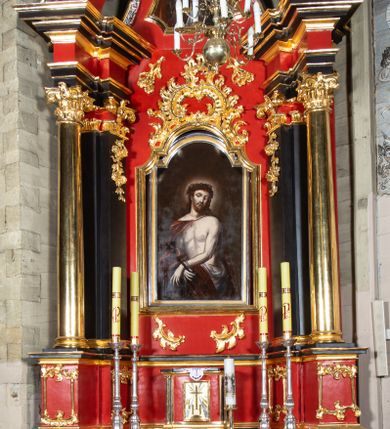 Zdjęcie nr 1: Nastawa architektoniczna, jednokondygnacyjna, jednoosiowa ze zwieńczeniem. Ołtarz drewniany, prostopadłościenny. Antepedium w kształcie leżącego prostokąta, ujęte profilowaną, polichromowaną, złoconą i srebrzoną ramą z ornamentyką roślinną. Pole wypełnione czerwoną tkaniną z haftem z gorejącym sercem oplecionym koroną cierniową pośrodku, flankowanym parą przeciwstawnie ustawionych gałązek białej lilii. Tabernakulum w kształcie prostopadłościennej skrzynki na niskim cokole, z lekko wypukłą ścianką przednią, flankowaną parą pilastrów z przylegającymi do nich półkolumienkami wspierającymi wyłamujący się ponad nimi gzyms, lekko wypukły w środkowej partii. Drzwiczki w kształcie stojącego prostokąta, ujęte profilowaną i złoconą ramą, zdobione grawerowanym, złoconym i srebrzonym przedstawieniem krzyża łacińskiego z wiązkami promieni wychodzących z miejsca przecięcia ramion oraz monogramem IHS. Nastawa ustawiona na zdwojonym, uskokowym cokole, ujęta parą filarów na rzucie kwadratu i stojącą na ich tle parą kolumn o gładkich trzonach i akantowo-wolutowych kapitelach, wspierających wyłamujące się ponad nimi, skośnie ustawione, przerwane belkowanie z silnie wysuniętym gzymsem. Zwieńczenie w formie kartusza o faliście ukształtowanych bokach, zamknięte przerwanym gzymsem w kształcie łuku wklęsło-wypukłego, flankowane przerwanym przyczółkiem ukształtowanym wolutowo, zakończone rocaille&#039;ami. Pole środkowe retabulum w dolnej i górnej partii zdobione ornamentyką rocaille&#039;ową oraz motywem gałązek różanych. W polu głównym nastawy obraz Ecce Homo, a w zwieńczeniu obraz św. Jana Nepomucena. Struktura polichromowana w kolorze czerwieni i czerni, detal architektoniczny i ornamentyka złocone oraz srebrzone. Według dokumentacji konserwatorskiej z 2014 roku na trzonie kolumny znajduje się napis „[...] Pozłotnicy, i z Warszawy emigrant”.