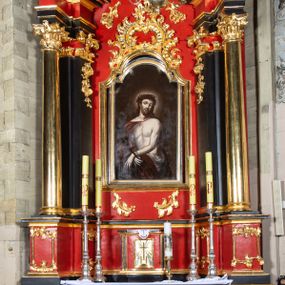 Zdjęcie nr 1: Nastawa architektoniczna, jednokondygnacyjna, jednoosiowa ze zwieńczeniem. Ołtarz drewniany, prostopadłościenny. Antepedium w kształcie leżącego prostokąta, ujęte profilowaną, polichromowaną, złoconą i srebrzoną ramą z ornamentyką roślinną. Pole wypełnione czerwoną tkaniną z haftem z gorejącym sercem oplecionym koroną cierniową pośrodku, flankowanym parą przeciwstawnie ustawionych gałązek białej lilii. Tabernakulum w kształcie prostopadłościennej skrzynki na niskim cokole, z lekko wypukłą ścianką przednią, flankowaną parą pilastrów z przylegającymi do nich półkolumienkami wspierającymi wyłamujący się ponad nimi gzyms, lekko wypukły w środkowej partii. Drzwiczki w kształcie stojącego prostokąta, ujęte profilowaną i złoconą ramą, zdobione grawerowanym, złoconym i srebrzonym przedstawieniem krzyża łacińskiego z wiązkami promieni wychodzących z miejsca przecięcia ramion oraz monogramem IHS. Nastawa ustawiona na zdwojonym, uskokowym cokole, ujęta parą filarów na rzucie kwadratu i stojącą na ich tle parą kolumn o gładkich trzonach i akantowo-wolutowych kapitelach, wspierających wyłamujące się ponad nimi, skośnie ustawione, przerwane belkowanie z silnie wysuniętym gzymsem. Zwieńczenie w formie kartusza o faliście ukształtowanych bokach, zamknięte przerwanym gzymsem w kształcie łuku wklęsło-wypukłego, flankowane przerwanym przyczółkiem ukształtowanym wolutowo, zakończone rocaille'ami. Pole środkowe retabulum w dolnej i górnej partii zdobione ornamentyką rocaille'ową oraz motywem gałązek różanych. W polu głównym nastawy obraz Ecce Homo, a w zwieńczeniu obraz św. Jana Nepomucena. Struktura polichromowana w kolorze czerwieni i czerni, detal architektoniczny i ornamentyka złocone oraz srebrzone. Według dokumentacji konserwatorskiej z 2014 roku na trzonie kolumny znajduje się napis „[...] Pozłotnicy, i z Warszawy emigrant”.