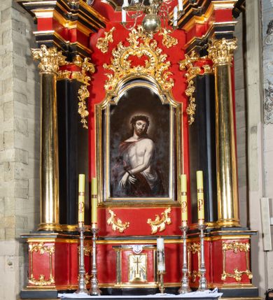 Zdjęcie nr 1: Tabernakulum w kształcie prostopadłościennej skrzynki na niskim cokole, z lekko wypukłą ścianką przednią, flankowaną parą pilastrów z przylegającymi do nich półkolumienkami wspierającymi wyłamujący się ponad nimi gzyms, lekko wypukły w środkowej partii. Drzwiczki w kształcie stojącego prostokąta, ujęte profilowaną i złoconą ramą, zdobione grawerowanym, złoconym i srebrzonym przedstawieniem krzyża łacińskiego z wiązkami promieni wychodzących z miejsca przecięcia ramion oraz monogramem IHS. Struktura polichromowana w kolorze czerwieni i czerni, partie detalu architektonicznego złocone.   
