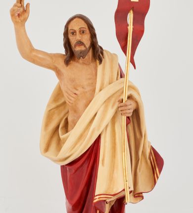 Zdjęcie nr 1: Rzeźba Chrystusa Zmartwychwstałego pełnoplastyczna, wolnostojąca. Chrystus ukazany jest w całej postaci, ustawiony frontalnie, w delikatnym kontrapoście; prawą, wysoko uniesioną ręką czyni gest błogosławieństwa, a w lewej trzyma chorągiew o długim drzewcu zakończonym krzyżem, na czerwonej tkaninie widnieje złoty krzyż. Chrystus ma podłużną twarz z długim i wąskim nosem, wzrokiem skierowanym w dal, okoloną krótką i ciemną brodą, rozdzieloną na końcu w dwa pukle oraz długie, ciemnobrązowe włosy, które opadają na plecy i ramiona. Chrystus jest ubrany w czerwony płaszcz z jasnobeżową podszewką i złoconą bordiurą, przerzucony przez lewe ramię i odsłaniający prawe ramię oraz nagi tors z drugą połą płaszcza zarzuconą na lewą rękę. Na dłoniach, stopach i boku zaznaczone są ślady męki. Polichromia ciała jest naturalistyczna. Rzeźba ustawiona jest na dwóch ukośnie ustawionych, prostopadłościennych kostkach. 