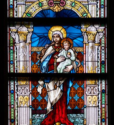 Zdjęcie nr 1: Witraż w kształcie stojącego prostokąta, zamkniętego łukiem odcinkowym. W centrum kompozycji ukazana jest Matka Boska z Dzieciątkiem wewnątrz dekoracyjnej aediculi. Maria przedstawiona jest frontalnie, na lewej ręce trzyma Dzieciątko Jezus, a prawą je podtrzymuje. Twarz podłużna o ciemnej karnacji z wysokim czołem, długim nosem, dużymi oczami i małymi ustami. Maria ubrana jest w czerwoną suknię; biały płaszcz z niebieską podszewką, spięty na dekolcie romboidalną klamrą oraz biały welon na głowie, spod którego spływają jej bujne i długie włosy. Dzieciątko ukazane jest w pozycji siedzącej, zwrócone trzy czwarte w prawo, w prawej dłoni trzyma różaniec, a w lewej złote jabłko. Dzieciątko ubrane jest w jasnozieloną suknię z długimi rękawami. Ma podłużną twarz o jasnej karnacji z dużymi oczami i długim nosem, okoloną kędzierzawymi, jasnymi włosami. Maria i Dzieciątko mają wokół głów koliste nimby. 

Aedicula wsparta jest na parach kolumn oraz stojących za nimi filarów, ustawionych na dwustopniowych cokołach. Podpory podtrzymują zwieńczenie z łukiem odcinkowym z przodu. W tle aediculi na prostym drążku, zawieszona jest kotara dekorowana rzędami złotych medalionów wypełnionych liliami andegaweńskimi oraz znajdującymi się między nimi niebieskimi rozetkami umieszczonymi w romboidalnych obramieniach. Cokoły kolumn dekorowane są płycinami w kształcie leżących prostokątów o wciętych narożach. Kolumny stoją na złoconych bazach, trzony są w jednej trzeciej wysokości kanelowane i dekorowane liśćmi akantu, przedzielone szeroką opaską z wolutami, wyżej gładkie, zdobione u dołu akantem, a u góry podwieszonymi festonami; kapitele kolumn zdobione liśćmi akantu. Zwieńczenie dwustopniowe, po bokach flankowane filarkami bogato dekorowane wolutkami i liśćmi akantu, na szczycie trójkątny przyczółek. W dolnej partii witrażu, na cokole aediculi kartusz zdobiony stylizowanym ornamentem okuciowym z napisem w polu: „SANCTA MARIA!” oraz sygnatura artysty „F(riedrich) DORN u.[?] C2. MUENCEHN.”. Tło dekorowane rozetkami w kwadratowych przeszkleniach. Całość obiega pas dekoracyjnej bordiury utworzonej z rzędu campanul i rozetek.
