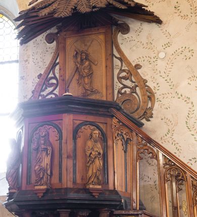 Zdjęcie nr 2: Ambona przyścienna, koszowa, z dostępem od zachodu w postaci jednobiegowych schodów z ażurową dekorowaną stylizowanymi blendami balustradą. Oparty na słupie w formie pnia drzewa, które korzeniami wchodzi w trójstopniową podstawę kosz na planie ściętego ośmioboku. Pola ścianek w formie płycin zamkniętych łukiem półkolistym z motywem fryzu arkadkowego. Przed każdą z płycin, ustawiona na konsolce całopostaciowa figurka jednego z czterech ewangelistów trzymających w dłoniach atrybuty (św. Mateusz z księgą i piórem, św. Łukasz ze zwojem i stylusem, św. Jan z kielichem z wężem, św. Marek z księgą). Podstawę kosza obiega gzyms, podwieszenie ujmują konary w promienistym układzie, pomiędzy którymi są symbole ewangelistów: anioł, wół, orzeł i lew. Parapet w formie wklęsło-wypukłego gzymsu. Zaplecek w kształcie stojącego prostokąta, ujętego ramą oraz parą ażurowych wolut; w polu arkada z płaskorzeźbionym przedstawieniem Chrystusa niosącego krzyż, w szczycie napis IHS ujęty wiązkami promienistej glorii. Baldachim utworzony z palmowych liści. Na podniebiu gołębicą Ducha Świętego w glorii promienistej.