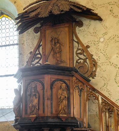 Zdjęcie nr 1: Ambona przyścienna, koszowa, z dostępem od zachodu w postaci jednobiegowych schodów z ażurową dekorowaną stylizowanymi blendami balustradą. Oparty na słupie w formie pnia drzewa, które korzeniami wchodzi w trójstopniową podstawę kosz na planie ściętego ośmioboku. Pola ścianek w formie płycin zamkniętych łukiem półkolistym z motywem fryzu arkadkowego. Przed każdą z płycin, ustawiona na konsolce całopostaciowa figurka jednego z czterech ewangelistów trzymających w dłoniach atrybuty (św. Mateusz z księgą i piórem, św. Łukasz ze zwojem i stylusem, św. Jan z kielichem z wężem, św. Marek z księgą). Podstawę kosza obiega gzyms, podwieszenie ujmują konary w promienistym układzie, pomiędzy którymi są symbole ewangelistów: anioł, wół, orzeł i lew. Parapet w formie wklęsło-wypukłego gzymsu. Zaplecek w kształcie stojącego prostokąta, ujętego ramą oraz parą ażurowych wolut; w polu arkada z płaskorzeźbionym przedstawieniem Chrystusa niosącego krzyż, w szczycie napis IHS ujęty wiązkami promienistej glorii. Baldachim utworzony z palmowych liści. Na podniebiu gołębicą Ducha Świętego w glorii promienistej.