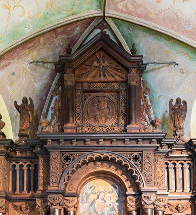 Zdjęcie nr 2: Ołtarz architektoniczny, jednokondygnacyjny, trójosiowy, ze zwieńczeniem. Ołtarz prostopadłościenny, kamienny, ustawiony na podstawie, w drewnianej obudowie. Mensa drewniana z marmurowym portatylem. Tabernakulum w formie prostopadłościennej metalowej skrzynki w drewnianej obudowie. Metalowe drzwiczki zdobi motyw kielicha eucharystycznego. Tabernakulum ujęte jest po bokach parą wolnostojących kolumienek wspierających wysunięte ponad nimi, wieńczące je belkowanie z wysokim fryzem. W partii belkowania nad drzwiczkami arkada dekorowana koronką i maswerkowymi rozetami w przyłuczach. Na belkowaniu tron wystawienia, na którym ustawiony krucyfiks. Osadzona na cokole nastawa ołtarza ma formę trójprzelotowego łuku triumfalnego. Część środkowa w kształcie półkolistej arkady wspartej na dwóch parach kolumn, z których wewnętrzne są cofnięte względem zewnętrznych. Arkady artykułujące osie boczne są mniejsze, ujmuje je po jednej parze kolumn wspierających zryzalitowane cokoły flankujące pola wypełnione fryzem arkadkowym. Zwieńczenie ołtarza ujmuje para kolumn wspierających gzyms w formie trójkątnego przyczółka. Pola cokołów retabulum zdobi stylizowana ornamentyka roślinna, kapitele kolumn oraz zwieńczenia arkad osi bocznych motywy uskrzydlonych główek anielskich. Krawędź łuku arkady części środkowej obiega fryz arkadkowy. W przyłuczach arkady ażurowe maswerki. Gzyms wieńczący kondygnację retabulum ujmuje motyw konsolek w formie liści akantu. Zwieńczenie flankowane parą ażurowych uszaków w postaci ornamentu roślinnego, parą figur aniołów oraz dwiema parami wazonów. Pole przyczółka wypełnia płaskorzeźbione przedstawienie gołębicy Ducha Świętego na tle promienistej glorii. Po jego bokach znajduje się para wazonów, szczyt jest w formie krzyża. Zwieńczenie flankują dwie figury aniołów ujęte po bokach dwoma parami wazonów.  
Pole części środkowej w kształcie stojącego prostokąta zamkniętego półkoliście z obrazem Madonny z Dzieciątkiem. W niszach osi bocznych są figury św. Piotra po prawej stronie i św. Pawła po lewej. Kształt pola zwieńczenia jest zbliżony do kwadratu ujętego szerokim obramieniem zdobionym parami S-kształtnych wolut. W środku znajduje się tondo z płaskorzeźbionym przedstawieniem anioła trzymającego kwiat lilii. 

Struktura retabulum utrzymana w kolorze drewna, trzony kolumn i część detali architektonicznych marmoryzowane, tła osi bocznych polichromowane w kolorze ciemnoniebieskim.    

