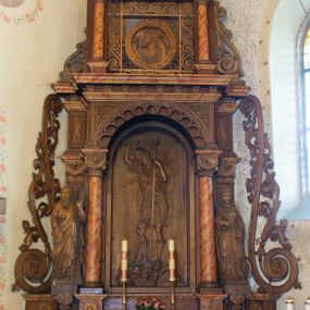 Zdjęcie nr 1: Ołtarz architektoniczny, jednokondygnacyjny, jednoosiowy ze zwieńczeniem. Ołtarz prostopadłościenny z drewnianą mensą, po bokach ujęty uskokowo. Retabulum osadzone na niewysokim, podwójnie zryzalitowanym po bokach cokole, skrajne partie ustawione ukośnie. Część środkowa w formie półkoliście zamkniętej arkady wspartej na parze kolumn i ujętej parą pilastrów. Całość wieńczy wyłamujące się w części środkowej belkowanie. Zwieńczenie ujmuje para kolumn osadzonych na niewielkich cokołach i wspierających gzyms w kształcie trójkątnego przyczółka. Pola uskokowych boków ołtarza zdobią motywy S-kształtnych, ulistnionych wolut, pole antepedium wypełnia fryz arkadkowy. Partie cokołu retabulum zdobią motywy rozet. W kapitelach kolumn jest dekoracja w formie uskrzydlonych główek anielskich. Łuk arkady zdobi ażurowy fryz arkadkowy, natomiast górne pola tej części retabulum motywy czwórliści i S-kształtnych, ulistnionych wolut. Zarówno środkową część retabulum, jak i zwieńczenie ujmują po bokach pary ażurowych uszaków w formie wici roślinnej. W polu przyczółka jest płaskorzeźbiona promienista gloria z monogramem maryjnym pośrodku. Szczyt zwieńczenia w formie krzyża. Pole arkady części środkowej retabulum wypełnia płaskorzeźbiona sylwetka św. Michała Archanioła, natomiast po bokach, na tle pilastrów została ustawiona para figur św. Katarzyny Aleksandryjskiej po prawej stronie i św. Andrzeja po lewej. Pole zwieńczenia w kształcie zbliżonym do kwadratu, ujęte szerokim obramieniem zdobionym ażurowym ornamentem w formie S-kształtnych, ulistnionych wolut. W środku jest tondo z płaskorzeźbionym wizerunkiem św. Jana Chrzciciela. 
Struktura retabulum utrzymana w kolorze drewna, trzony kolumn i część detali architektonicznych marmoryzowane.         
