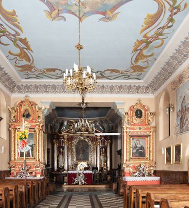 Zdjęcie nr 17: Kościół orientowany, murowany, jednonawowy, z trójbocznie zamkniętym prezbiterium z zakrystią od północy i kruchtą przylegającą do nawy od południa. Prezbiterium jest niższe i węższe od nawy, obie części mają gładkie ściany przeprute jedynie oknami w kształcie stojącego prostokąta zamkniętego łukiem półkolistym – cztery w prezbiterium, sześć w nawie. Sklepienia obu części zwierciadlane. Od południa chór o prostej balustradzie, wsparty na dwóch kolumnach. Ściany pokrywa polichromia ze scenami i cytatami z Pisma Świętego, postaciami świętych oraz żyjących, a także o motywach dekoracyjnych – roślinnych. Do około jednej czwartej wysokości ścian okładzina.  Zachodnią elewację frontową kościoła tworzy trójosiowa, wieżowa fasada. Oś kompozycji stanowi wysunięta ryzalitowo czterokondygnacyjna wieża. Ściana czołowa wieży dekorowana w pierwszej i drugiej kondygnacji boniowaniem i oknami w kształcie leżącego prostokąta osłoniętymi ozdobną kratą, z portalem o wykroju prostokątnym, ponad którym napis „DEO SOLI GLORIA”. W drugiej i trzeciej kondygnacji okna o wykroju stojącego prostokąta zamkniętego łukiem pełnym. Niżej w płycinie zwieńczonej łukiem nadwieszonym, wyżej w prostokątnej. Kondygnacje rozdzielone za pomocą masywnych gzymsów. Całość zwieńczona dachem namiotowym z kulą i krzyżem. Osie boczne cofnięte w stosunku do wysuniętej wieży i zrytmizowane podziałem ramowym, wypełnione wklęsłą płyciną w formie wydłużonego, stojącego prostokąta. Nad gzymsem fragment przyczółka przysłonięty wieżą. Elewacje boczne i tylna ukształtowane jednorodnie, gładkie, przeprute jedynie oknami. Wszystkie dachy blaszane – nad nawą i kruchtą dwuspadowy, nad zakrystią pulpitowy, nad prezbiterium zamknięty wielobocznie. 
Wartymi uwagi elementami wyposażenia kościoła jest siedemnastowieczny ołtarz boczny prawy z późniejszymi o wiek wizerunkami św. Katarzyny i św. Wawrzyńca, bogato rzeźbiona ława kolatorska i siedemnastowieczna chrzcielnica. 