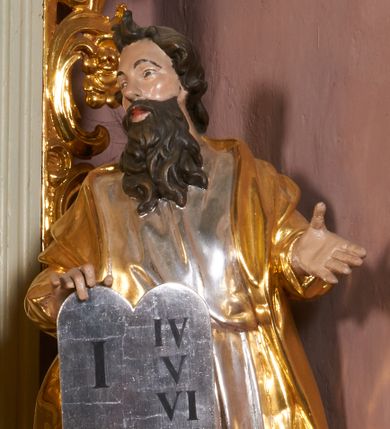 Zdjęcie nr 1: Rzeźba jest drążona z tyłu, ustawiona na niewielkiej, prostej podstawie. Mojżesz przedstawiony jest w całej postaci, ukazany frontalnie, stoi w kontrapoście, z głową skierowaną w prawą stronę. Prawą dłonią trzyma tablice z przykazaniami, częściowo przysłaniające przód postaci, lewą wyciąga przed siebie. Twarz ma szczupłą i pociągłą, o przymrużonych oczach i uniesionych brwiach, prostym nosie i pełnych, rozchylonych ustach, okoloną długą, kręconą brodą opadającą na pierś oraz krótkimi, zmierzwionymi włosami, na szczycie głowy uformowanymi w dwa loki. Mojżesz ubrany jest w tunikę, przepasaną w talii oraz płaszcz z rękawami narzucony na plecy i ramiona. Figura polichromowana naturalistycznie w partiach ciała, tunika srebrzona, płaszcz złocony.