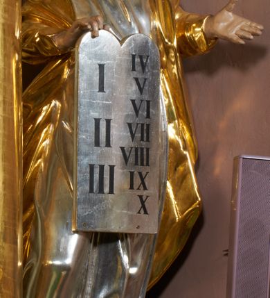 Zdjęcie nr 2: Rzeźba jest drążona z tyłu, ustawiona na niewielkiej, prostej podstawie. Mojżesz przedstawiony jest w całej postaci, ukazany frontalnie, stoi w kontrapoście, z głową skierowaną w prawą stronę. Prawą dłonią trzyma tablice z przykazaniami, częściowo przysłaniające przód postaci, lewą wyciąga przed siebie. Twarz ma szczupłą i pociągłą, o przymrużonych oczach i uniesionych brwiach, prostym nosie i pełnych, rozchylonych ustach, okoloną długą, kręconą brodą opadającą na pierś oraz krótkimi, zmierzwionymi włosami, na szczycie głowy uformowanymi w dwa loki. Mojżesz ubrany jest w tunikę, przepasaną w talii oraz płaszcz z rękawami narzucony na plecy i ramiona. Figura polichromowana naturalistycznie w partiach ciała, tunika srebrzona, płaszcz złocony.