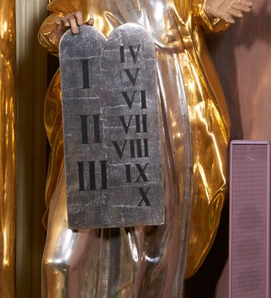 Zdjęcie nr 3: Rzeźba jest drążona z tyłu, ustawiona na niewielkiej, prostej podstawie. Mojżesz przedstawiony jest w całej postaci, ukazany frontalnie, stoi w kontrapoście, z głową skierowaną w prawą stronę. Prawą dłonią trzyma tablice z przykazaniami, częściowo przysłaniające przód postaci, lewą wyciąga przed siebie. Twarz ma szczupłą i pociągłą, o przymrużonych oczach i uniesionych brwiach, prostym nosie i pełnych, rozchylonych ustach, okoloną długą, kręconą brodą opadającą na pierś oraz krótkimi, zmierzwionymi włosami, na szczycie głowy uformowanymi w dwa loki. Mojżesz ubrany jest w tunikę, przepasaną w talii oraz płaszcz z rękawami narzucony na plecy i ramiona. Figura polichromowana naturalistycznie w partiach ciała, tunika srebrzona, płaszcz złocony.