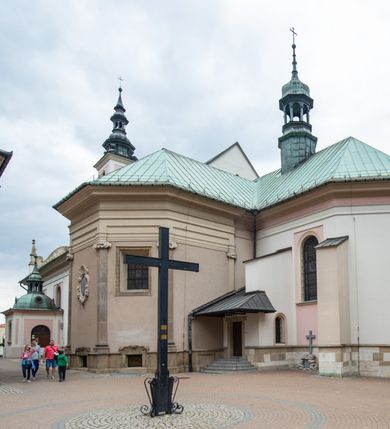 Zdjęcie nr 18: Kościół orientowany, murowany, trzynawowy. Typ halowy, z węższym, prostokątnym prezbiterium zamkniętym trójbocznie oraz z wieżą wbudowaną w zachodnią fasadę. Do prezbiterium od północy przylega zakrystia (do niej od zachodu klatka schodowa wiodąca do skarbca na piętrze), od południa natomiast kaplica Morsztynów z XVII wieku. Kaplica na planie prostokąta zamkniętego trójbocznie, z niszami w ściętych narożach, pod nią sklepiona krypta grobowa, opięta pilastrami wewnątrz i na zewnątrz. Od południa do korpusu przylega kruchta stanowiąca również kaplicę Matki Boskiej Pocieszenia. Korpus czteroprzęsłowy, nawy oddzielają arkady wsparte na filarach, pierwsze stanowią podparcie dla chóru. Nad nawami i prezbiterium strop imitujący sklepienie, na którym malowana dekoracja imitująca gurty, plafony oraz stiuki. Dekoracja ścian powtarza podziały wyznaczone przez arkady, namalowano na nich pilastry podtrzymujące belkowanie. W prezbiterium dwa okna usytuowane niedaleko ołtarza, w nawie osiem, wszystkie w kształcie stojącego prostokąta zamkniętego półkoliście, wypełnione witrażami z dekoracją geometryczną. Główne wejście od zachodu, drugie od północy, trzecie symetrycznie od południa przez kruchtę sklepioną kopułą, kolejne trzy do prezbiterium, od południa przez zakrystię i od północy przez kruchtę. 
Fasada zachodnia na cokole, podzielona gzymsami i lizenami, z wieżą zakończoną hełmem. Na osi drzwi w kształcie stojącego prostokąta flankowane prostokątnymi oknami, powyżej trzecie, większe okno zamknięte łukiem odcinkowym; kondygnację wieńczy gzyms. Druga kondygnacja powtarza podziały pierwszej. W środkowej osi okulus, powyżej dwudzielne okno zamknięte półkoliście. Nad nim tarcza zegara, kolejne dwie na ścianach bocznych. Kondygnacja ograniczona po bokach spływali wolutowymi i obeliskami zakończonymi kulami; przechodzi w wieżę. Powyżej gzyms podtrzymywany kroksztynami. Ostatnia kondygnacja o artykulacji ramowej z balkonami z każdej strony, powyżej wieńczącego gzymsu. Pozostałe elewacje na cokole, kontynuują podziały  lizenowo-ramowe, artykulacją podkreślają okna. Kaplica Morsztynów opięta pilastrami z głowicami, prezbiterium oszkarpowane. Dach nad nawą główną, kaplicą i prezbiterium dwuspadowy, na kruchtą południową kopuła, na wieży ozdobny hełm dołem wklęsły, dekorowany wolutami. Powyżej kopuła, na której latarnia przepruta arkadami i zwieńczona przewężoną w środku kopułą zakończoną ostrosłupem z kulą i krzyżem. 
Do najcenniejszych zabytków świątyni należą m.in. zaprojektowana przez Jana Delamarsa kaplica Morsztynów dekorowana stiukami z 1693 roku autorstwa Baltazara Fontany. Wewnątrz niej warto zwrócić uwagę na osiemnastowieczny ołtarz z przedstawieniem Oblicza Chrystusa i rzeźbionym antepedium. W ołtarz Ukrzyżowania znajdujący się w północnej nawie kościoła wkomponowano gotyckie rzeźby św. Jana i Marii. 