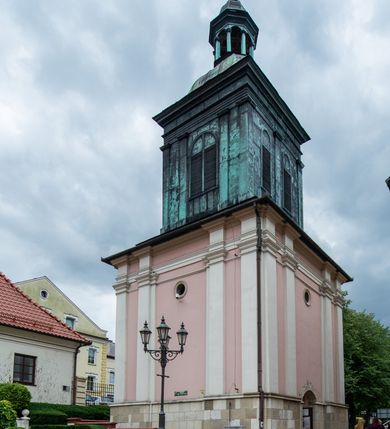 Zdjęcie nr 48: Kościół orientowany, murowany, trzynawowy. Typ halowy, z węższym, prostokątnym prezbiterium zamkniętym trójbocznie oraz z wieżą wbudowaną w zachodnią fasadę. Do prezbiterium od północy przylega zakrystia (do niej od zachodu klatka schodowa wiodąca do skarbca na piętrze), od południa natomiast kaplica Morsztynów z XVII wieku. Kaplica na planie prostokąta zamkniętego trójbocznie, z niszami w ściętych narożach, pod nią sklepiona krypta grobowa, opięta pilastrami wewnątrz i na zewnątrz. Od południa do korpusu przylega kruchta stanowiąca również kaplicę Matki Boskiej Pocieszenia. Korpus czteroprzęsłowy, nawy oddzielają arkady wsparte na filarach, pierwsze stanowią podparcie dla chóru. Nad nawami i prezbiterium strop imitujący sklepienie, na którym malowana dekoracja imitująca gurty, plafony oraz stiuki. Dekoracja ścian powtarza podziały wyznaczone przez arkady, namalowano na nich pilastry podtrzymujące belkowanie. W prezbiterium dwa okna usytuowane niedaleko ołtarza, w nawie osiem, wszystkie w kształcie stojącego prostokąta zamkniętego półkoliście, wypełnione witrażami z dekoracją geometryczną. Główne wejście od zachodu, drugie od północy, trzecie symetrycznie od południa przez kruchtę sklepioną kopułą, kolejne trzy do prezbiterium, od południa przez zakrystię i od północy przez kruchtę. 
Fasada zachodnia na cokole, podzielona gzymsami i lizenami, z wieżą zakończoną hełmem. Na osi drzwi w kształcie stojącego prostokąta flankowane prostokątnymi oknami, powyżej trzecie, większe okno zamknięte łukiem odcinkowym; kondygnację wieńczy gzyms. Druga kondygnacja powtarza podziały pierwszej. W środkowej osi okulus, powyżej dwudzielne okno zamknięte półkoliście. Nad nim tarcza zegara, kolejne dwie na ścianach bocznych. Kondygnacja ograniczona po bokach spływali wolutowymi i obeliskami zakończonymi kulami; przechodzi w wieżę. Powyżej gzyms podtrzymywany kroksztynami. Ostatnia kondygnacja o artykulacji ramowej z balkonami z każdej strony, powyżej wieńczącego gzymsu. Pozostałe elewacje na cokole, kontynuują podziały  lizenowo-ramowe, artykulacją podkreślają okna. Kaplica Morsztynów opięta pilastrami z głowicami, prezbiterium oszkarpowane. Dach nad nawą główną, kaplicą i prezbiterium dwuspadowy, na kruchtą południową kopuła, na wieży ozdobny hełm dołem wklęsły, dekorowany wolutami. Powyżej kopuła, na której latarnia przepruta arkadami i zwieńczona przewężoną w środku kopułą zakończoną ostrosłupem z kulą i krzyżem. 
Do najcenniejszych zabytków świątyni należą m.in. zaprojektowana przez Jana Delamarsa kaplica Morsztynów dekorowana stiukami z 1693 roku autorstwa Baltazara Fontany. Wewnątrz niej warto zwrócić uwagę na osiemnastowieczny ołtarz z przedstawieniem Oblicza Chrystusa i rzeźbionym antepedium. W ołtarz Ukrzyżowania znajdujący się w północnej nawie kościoła wkomponowano gotyckie rzeźby św. Jana i Marii. 