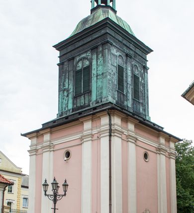 Zdjęcie nr 35: Kościół orientowany, murowany, trzynawowy. Typ halowy, z węższym, prostokątnym prezbiterium zamkniętym trójbocznie oraz z wieżą wbudowaną w zachodnią fasadę. Do prezbiterium od północy przylega zakrystia (do niej od zachodu klatka schodowa wiodąca do skarbca na piętrze), od południa natomiast kaplica Morsztynów z XVII wieku. Kaplica na planie prostokąta zamkniętego trójbocznie, z niszami w ściętych narożach, pod nią sklepiona krypta grobowa, opięta pilastrami wewnątrz i na zewnątrz. Od południa do korpusu przylega kruchta stanowiąca również kaplicę Matki Boskiej Pocieszenia. Korpus czteroprzęsłowy, nawy oddzielają arkady wsparte na filarach, pierwsze stanowią podparcie dla chóru. Nad nawami i prezbiterium strop imitujący sklepienie, na którym malowana dekoracja imitująca gurty, plafony oraz stiuki. Dekoracja ścian powtarza podziały wyznaczone przez arkady, namalowano na nich pilastry podtrzymujące belkowanie. W prezbiterium dwa okna usytuowane niedaleko ołtarza, w nawie osiem, wszystkie w kształcie stojącego prostokąta zamkniętego półkoliście, wypełnione witrażami z dekoracją geometryczną. Główne wejście od zachodu, drugie od północy, trzecie symetrycznie od południa przez kruchtę sklepioną kopułą, kolejne trzy do prezbiterium, od południa przez zakrystię i od północy przez kruchtę. 
Fasada zachodnia na cokole, podzielona gzymsami i lizenami, z wieżą zakończoną hełmem. Na osi drzwi w kształcie stojącego prostokąta flankowane prostokątnymi oknami, powyżej trzecie, większe okno zamknięte łukiem odcinkowym; kondygnację wieńczy gzyms. Druga kondygnacja powtarza podziały pierwszej. W środkowej osi okulus, powyżej dwudzielne okno zamknięte półkoliście. Nad nim tarcza zegara, kolejne dwie na ścianach bocznych. Kondygnacja ograniczona po bokach spływali wolutowymi i obeliskami zakończonymi kulami; przechodzi w wieżę. Powyżej gzyms podtrzymywany kroksztynami. Ostatnia kondygnacja o artykulacji ramowej z balkonami z każdej strony, powyżej wieńczącego gzymsu. Pozostałe elewacje na cokole, kontynuują podziały  lizenowo-ramowe, artykulacją podkreślają okna. Kaplica Morsztynów opięta pilastrami z głowicami, prezbiterium oszkarpowane. Dach nad nawą główną, kaplicą i prezbiterium dwuspadowy, na kruchtą południową kopuła, na wieży ozdobny hełm dołem wklęsły, dekorowany wolutami. Powyżej kopuła, na której latarnia przepruta arkadami i zwieńczona przewężoną w środku kopułą zakończoną ostrosłupem z kulą i krzyżem. 
Do najcenniejszych zabytków świątyni należą m.in. zaprojektowana przez Jana Delamarsa kaplica Morsztynów dekorowana stiukami z 1693 roku autorstwa Baltazara Fontany. Wewnątrz niej warto zwrócić uwagę na osiemnastowieczny ołtarz z przedstawieniem Oblicza Chrystusa i rzeźbionym antepedium. W ołtarz Ukrzyżowania znajdujący się w północnej nawie kościoła wkomponowano gotyckie rzeźby św. Jana i Marii. 