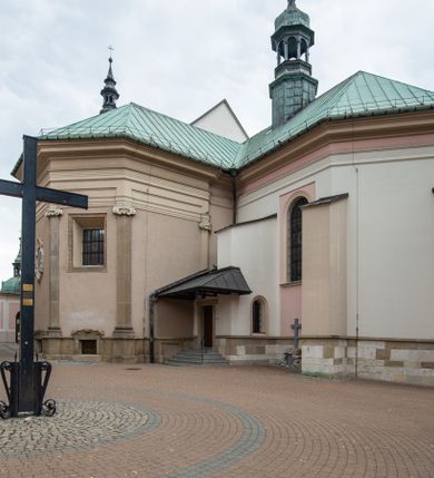 Zdjęcie nr 16: Kościół orientowany, murowany, trzynawowy. Typ halowy, z węższym, prostokątnym prezbiterium zamkniętym trójbocznie oraz z wieżą wbudowaną w zachodnią fasadę. Do prezbiterium od północy przylega zakrystia (do niej od zachodu klatka schodowa wiodąca do skarbca na piętrze), od południa natomiast kaplica Morsztynów z XVII wieku. Kaplica na planie prostokąta zamkniętego trójbocznie, z niszami w ściętych narożach, pod nią sklepiona krypta grobowa, opięta pilastrami wewnątrz i na zewnątrz. Od południa do korpusu przylega kruchta stanowiąca również kaplicę Matki Boskiej Pocieszenia. Korpus czteroprzęsłowy, nawy oddzielają arkady wsparte na filarach, pierwsze stanowią podparcie dla chóru. Nad nawami i prezbiterium strop imitujący sklepienie, na którym malowana dekoracja imitująca gurty, plafony oraz stiuki. Dekoracja ścian powtarza podziały wyznaczone przez arkady, namalowano na nich pilastry podtrzymujące belkowanie. W prezbiterium dwa okna usytuowane niedaleko ołtarza, w nawie osiem, wszystkie w kształcie stojącego prostokąta zamkniętego półkoliście, wypełnione witrażami z dekoracją geometryczną. Główne wejście od zachodu, drugie od północy, trzecie symetrycznie od południa przez kruchtę sklepioną kopułą, kolejne trzy do prezbiterium, od południa przez zakrystię i od północy przez kruchtę. 
Fasada zachodnia na cokole, podzielona gzymsami i lizenami, z wieżą zakończoną hełmem. Na osi drzwi w kształcie stojącego prostokąta flankowane prostokątnymi oknami, powyżej trzecie, większe okno zamknięte łukiem odcinkowym; kondygnację wieńczy gzyms. Druga kondygnacja powtarza podziały pierwszej. W środkowej osi okulus, powyżej dwudzielne okno zamknięte półkoliście. Nad nim tarcza zegara, kolejne dwie na ścianach bocznych. Kondygnacja ograniczona po bokach spływali wolutowymi i obeliskami zakończonymi kulami; przechodzi w wieżę. Powyżej gzyms podtrzymywany kroksztynami. Ostatnia kondygnacja o artykulacji ramowej z balkonami z każdej strony, powyżej wieńczącego gzymsu. Pozostałe elewacje na cokole, kontynuują podziały  lizenowo-ramowe, artykulacją podkreślają okna. Kaplica Morsztynów opięta pilastrami z głowicami, prezbiterium oszkarpowane. Dach nad nawą główną, kaplicą i prezbiterium dwuspadowy, na kruchtą południową kopuła, na wieży ozdobny hełm dołem wklęsły, dekorowany wolutami. Powyżej kopuła, na której latarnia przepruta arkadami i zwieńczona przewężoną w środku kopułą zakończoną ostrosłupem z kulą i krzyżem. 
Do najcenniejszych zabytków świątyni należą m.in. zaprojektowana przez Jana Delamarsa kaplica Morsztynów dekorowana stiukami z 1693 roku autorstwa Baltazara Fontany. Wewnątrz niej warto zwrócić uwagę na osiemnastowieczny ołtarz z przedstawieniem Oblicza Chrystusa i rzeźbionym antepedium. W ołtarz Ukrzyżowania znajdujący się w północnej nawie kościoła wkomponowano gotyckie rzeźby św. Jana i Marii. 