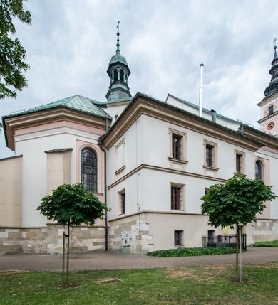 Zdjęcie nr 28: Kościół orientowany, murowany, trzynawowy. Typ halowy, z węższym, prostokątnym prezbiterium zamkniętym trójbocznie oraz z wieżą wbudowaną w zachodnią fasadę. Do prezbiterium od północy przylega zakrystia (do niej od zachodu klatka schodowa wiodąca do skarbca na piętrze), od południa natomiast kaplica Morsztynów z XVII wieku. Kaplica na planie prostokąta zamkniętego trójbocznie, z niszami w ściętych narożach, pod nią sklepiona krypta grobowa, opięta pilastrami wewnątrz i na zewnątrz. Od południa do korpusu przylega kruchta stanowiąca również kaplicę Matki Boskiej Pocieszenia. Korpus czteroprzęsłowy, nawy oddzielają arkady wsparte na filarach, pierwsze stanowią podparcie dla chóru. Nad nawami i prezbiterium strop imitujący sklepienie, na którym malowana dekoracja imitująca gurty, plafony oraz stiuki. Dekoracja ścian powtarza podziały wyznaczone przez arkady, namalowano na nich pilastry podtrzymujące belkowanie. W prezbiterium dwa okna usytuowane niedaleko ołtarza, w nawie osiem, wszystkie w kształcie stojącego prostokąta zamkniętego półkoliście, wypełnione witrażami z dekoracją geometryczną. Główne wejście od zachodu, drugie od północy, trzecie symetrycznie od południa przez kruchtę sklepioną kopułą, kolejne trzy do prezbiterium, od południa przez zakrystię i od północy przez kruchtę. 
Fasada zachodnia na cokole, podzielona gzymsami i lizenami, z wieżą zakończoną hełmem. Na osi drzwi w kształcie stojącego prostokąta flankowane prostokątnymi oknami, powyżej trzecie, większe okno zamknięte łukiem odcinkowym; kondygnację wieńczy gzyms. Druga kondygnacja powtarza podziały pierwszej. W środkowej osi okulus, powyżej dwudzielne okno zamknięte półkoliście. Nad nim tarcza zegara, kolejne dwie na ścianach bocznych. Kondygnacja ograniczona po bokach spływali wolutowymi i obeliskami zakończonymi kulami; przechodzi w wieżę. Powyżej gzyms podtrzymywany kroksztynami. Ostatnia kondygnacja o artykulacji ramowej z balkonami z każdej strony, powyżej wieńczącego gzymsu. Pozostałe elewacje na cokole, kontynuują podziały  lizenowo-ramowe, artykulacją podkreślają okna. Kaplica Morsztynów opięta pilastrami z głowicami, prezbiterium oszkarpowane. Dach nad nawą główną, kaplicą i prezbiterium dwuspadowy, na kruchtą południową kopuła, na wieży ozdobny hełm dołem wklęsły, dekorowany wolutami. Powyżej kopuła, na której latarnia przepruta arkadami i zwieńczona przewężoną w środku kopułą zakończoną ostrosłupem z kulą i krzyżem. 
Do najcenniejszych zabytków świątyni należą m.in. zaprojektowana przez Jana Delamarsa kaplica Morsztynów dekorowana stiukami z 1693 roku autorstwa Baltazara Fontany. Wewnątrz niej warto zwrócić uwagę na osiemnastowieczny ołtarz z przedstawieniem Oblicza Chrystusa i rzeźbionym antepedium. W ołtarz Ukrzyżowania znajdujący się w północnej nawie kościoła wkomponowano gotyckie rzeźby św. Jana i Marii. 