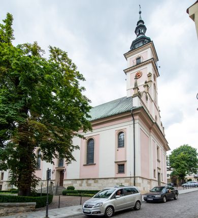 Zdjęcie nr 8: Kościół orientowany, murowany, trzynawowy. Typ halowy, z węższym, prostokątnym prezbiterium zamkniętym trójbocznie oraz z wieżą wbudowaną w zachodnią fasadę. Do prezbiterium od północy przylega zakrystia (do niej od zachodu klatka schodowa wiodąca do skarbca na piętrze), od południa natomiast kaplica Morsztynów z XVII wieku. Kaplica na planie prostokąta zamkniętego trójbocznie, z niszami w ściętych narożach, pod nią sklepiona krypta grobowa, opięta pilastrami wewnątrz i na zewnątrz. Od południa do korpusu przylega kruchta stanowiąca również kaplicę Matki Boskiej Pocieszenia. Korpus czteroprzęsłowy, nawy oddzielają arkady wsparte na filarach, pierwsze stanowią podparcie dla chóru. Nad nawami i prezbiterium strop imitujący sklepienie, na którym malowana dekoracja imitująca gurty, plafony oraz stiuki. Dekoracja ścian powtarza podziały wyznaczone przez arkady, namalowano na nich pilastry podtrzymujące belkowanie. W prezbiterium dwa okna usytuowane niedaleko ołtarza, w nawie osiem, wszystkie w kształcie stojącego prostokąta zamkniętego półkoliście, wypełnione witrażami z dekoracją geometryczną. Główne wejście od zachodu, drugie od północy, trzecie symetrycznie od południa przez kruchtę sklepioną kopułą, kolejne trzy do prezbiterium, od południa przez zakrystię i od północy przez kruchtę. 
Fasada zachodnia na cokole, podzielona gzymsami i lizenami, z wieżą zakończoną hełmem. Na osi drzwi w kształcie stojącego prostokąta flankowane prostokątnymi oknami, powyżej trzecie, większe okno zamknięte łukiem odcinkowym; kondygnację wieńczy gzyms. Druga kondygnacja powtarza podziały pierwszej. W środkowej osi okulus, powyżej dwudzielne okno zamknięte półkoliście. Nad nim tarcza zegara, kolejne dwie na ścianach bocznych. Kondygnacja ograniczona po bokach spływali wolutowymi i obeliskami zakończonymi kulami; przechodzi w wieżę. Powyżej gzyms podtrzymywany kroksztynami. Ostatnia kondygnacja o artykulacji ramowej z balkonami z każdej strony, powyżej wieńczącego gzymsu. Pozostałe elewacje na cokole, kontynuują podziały  lizenowo-ramowe, artykulacją podkreślają okna. Kaplica Morsztynów opięta pilastrami z głowicami, prezbiterium oszkarpowane. Dach nad nawą główną, kaplicą i prezbiterium dwuspadowy, na kruchtą południową kopuła, na wieży ozdobny hełm dołem wklęsły, dekorowany wolutami. Powyżej kopuła, na której latarnia przepruta arkadami i zwieńczona przewężoną w środku kopułą zakończoną ostrosłupem z kulą i krzyżem. 
Do najcenniejszych zabytków świątyni należą m.in. zaprojektowana przez Jana Delamarsa kaplica Morsztynów dekorowana stiukami z 1693 roku autorstwa Baltazara Fontany. Wewnątrz niej warto zwrócić uwagę na osiemnastowieczny ołtarz z przedstawieniem Oblicza Chrystusa i rzeźbionym antepedium. W ołtarz Ukrzyżowania znajdujący się w północnej nawie kościoła wkomponowano gotyckie rzeźby św. Jana i Marii. 