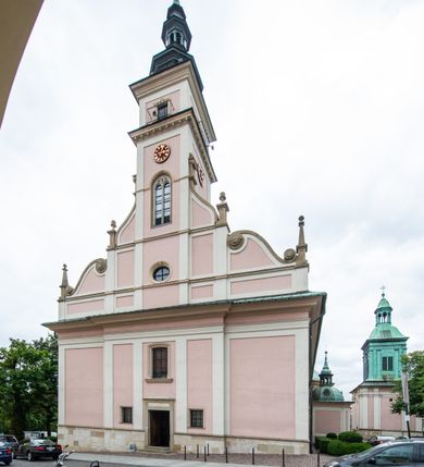 Zdjęcie nr 7: Kościół orientowany, murowany, trzynawowy. Typ halowy, z węższym, prostokątnym prezbiterium zamkniętym trójbocznie oraz z wieżą wbudowaną w zachodnią fasadę. Do prezbiterium od północy przylega zakrystia (do niej od zachodu klatka schodowa wiodąca do skarbca na piętrze), od południa natomiast kaplica Morsztynów z XVII wieku. Kaplica na planie prostokąta zamkniętego trójbocznie, z niszami w ściętych narożach, pod nią sklepiona krypta grobowa, opięta pilastrami wewnątrz i na zewnątrz. Od południa do korpusu przylega kruchta stanowiąca również kaplicę Matki Boskiej Pocieszenia. Korpus czteroprzęsłowy, nawy oddzielają arkady wsparte na filarach, pierwsze stanowią podparcie dla chóru. Nad nawami i prezbiterium strop imitujący sklepienie, na którym malowana dekoracja imitująca gurty, plafony oraz stiuki. Dekoracja ścian powtarza podziały wyznaczone przez arkady, namalowano na nich pilastry podtrzymujące belkowanie. W prezbiterium dwa okna usytuowane niedaleko ołtarza, w nawie osiem, wszystkie w kształcie stojącego prostokąta zamkniętego półkoliście, wypełnione witrażami z dekoracją geometryczną. Główne wejście od zachodu, drugie od północy, trzecie symetrycznie od południa przez kruchtę sklepioną kopułą, kolejne trzy do prezbiterium, od południa przez zakrystię i od północy przez kruchtę. 
Fasada zachodnia na cokole, podzielona gzymsami i lizenami, z wieżą zakończoną hełmem. Na osi drzwi w kształcie stojącego prostokąta flankowane prostokątnymi oknami, powyżej trzecie, większe okno zamknięte łukiem odcinkowym; kondygnację wieńczy gzyms. Druga kondygnacja powtarza podziały pierwszej. W środkowej osi okulus, powyżej dwudzielne okno zamknięte półkoliście. Nad nim tarcza zegara, kolejne dwie na ścianach bocznych. Kondygnacja ograniczona po bokach spływali wolutowymi i obeliskami zakończonymi kulami; przechodzi w wieżę. Powyżej gzyms podtrzymywany kroksztynami. Ostatnia kondygnacja o artykulacji ramowej z balkonami z każdej strony, powyżej wieńczącego gzymsu. Pozostałe elewacje na cokole, kontynuują podziały  lizenowo-ramowe, artykulacją podkreślają okna. Kaplica Morsztynów opięta pilastrami z głowicami, prezbiterium oszkarpowane. Dach nad nawą główną, kaplicą i prezbiterium dwuspadowy, na kruchtą południową kopuła, na wieży ozdobny hełm dołem wklęsły, dekorowany wolutami. Powyżej kopuła, na której latarnia przepruta arkadami i zwieńczona przewężoną w środku kopułą zakończoną ostrosłupem z kulą i krzyżem. 
Do najcenniejszych zabytków świątyni należą m.in. zaprojektowana przez Jana Delamarsa kaplica Morsztynów dekorowana stiukami z 1693 roku autorstwa Baltazara Fontany. Wewnątrz niej warto zwrócić uwagę na osiemnastowieczny ołtarz z przedstawieniem Oblicza Chrystusa i rzeźbionym antepedium. W ołtarz Ukrzyżowania znajdujący się w północnej nawie kościoła wkomponowano gotyckie rzeźby św. Jana i Marii. 