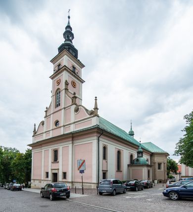 Zdjęcie nr 6: Kościół orientowany, murowany, trzynawowy. Typ halowy, z węższym, prostokątnym prezbiterium zamkniętym trójbocznie oraz z wieżą wbudowaną w zachodnią fasadę. Do prezbiterium od północy przylega zakrystia (do niej od zachodu klatka schodowa wiodąca do skarbca na piętrze), od południa natomiast kaplica Morsztynów z XVII wieku. Kaplica na planie prostokąta zamkniętego trójbocznie, z niszami w ściętych narożach, pod nią sklepiona krypta grobowa, opięta pilastrami wewnątrz i na zewnątrz. Od południa do korpusu przylega kruchta stanowiąca również kaplicę Matki Boskiej Pocieszenia. Korpus czteroprzęsłowy, nawy oddzielają arkady wsparte na filarach, pierwsze stanowią podparcie dla chóru. Nad nawami i prezbiterium strop imitujący sklepienie, na którym malowana dekoracja imitująca gurty, plafony oraz stiuki. Dekoracja ścian powtarza podziały wyznaczone przez arkady, namalowano na nich pilastry podtrzymujące belkowanie. W prezbiterium dwa okna usytuowane niedaleko ołtarza, w nawie osiem, wszystkie w kształcie stojącego prostokąta zamkniętego półkoliście, wypełnione witrażami z dekoracją geometryczną. Główne wejście od zachodu, drugie od północy, trzecie symetrycznie od południa przez kruchtę sklepioną kopułą, kolejne trzy do prezbiterium, od południa przez zakrystię i od północy przez kruchtę. 
Fasada zachodnia na cokole, podzielona gzymsami i lizenami, z wieżą zakończoną hełmem. Na osi drzwi w kształcie stojącego prostokąta flankowane prostokątnymi oknami, powyżej trzecie, większe okno zamknięte łukiem odcinkowym; kondygnację wieńczy gzyms. Druga kondygnacja powtarza podziały pierwszej. W środkowej osi okulus, powyżej dwudzielne okno zamknięte półkoliście. Nad nim tarcza zegara, kolejne dwie na ścianach bocznych. Kondygnacja ograniczona po bokach spływali wolutowymi i obeliskami zakończonymi kulami; przechodzi w wieżę. Powyżej gzyms podtrzymywany kroksztynami. Ostatnia kondygnacja o artykulacji ramowej z balkonami z każdej strony, powyżej wieńczącego gzymsu. Pozostałe elewacje na cokole, kontynuują podziały  lizenowo-ramowe, artykulacją podkreślają okna. Kaplica Morsztynów opięta pilastrami z głowicami, prezbiterium oszkarpowane. Dach nad nawą główną, kaplicą i prezbiterium dwuspadowy, na kruchtą południową kopuła, na wieży ozdobny hełm dołem wklęsły, dekorowany wolutami. Powyżej kopuła, na której latarnia przepruta arkadami i zwieńczona przewężoną w środku kopułą zakończoną ostrosłupem z kulą i krzyżem. 
Do najcenniejszych zabytków świątyni należą m.in. zaprojektowana przez Jana Delamarsa kaplica Morsztynów dekorowana stiukami z 1693 roku autorstwa Baltazara Fontany. Wewnątrz niej warto zwrócić uwagę na osiemnastowieczny ołtarz z przedstawieniem Oblicza Chrystusa i rzeźbionym antepedium. W ołtarz Ukrzyżowania znajdujący się w północnej nawie kościoła wkomponowano gotyckie rzeźby św. Jana i Marii. 