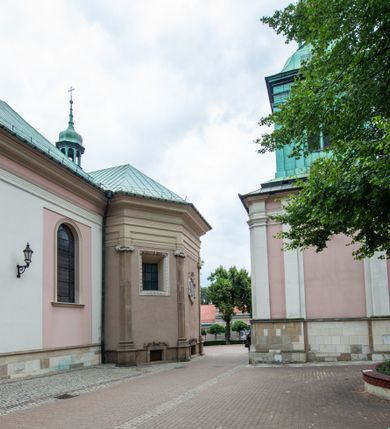 Zdjęcie nr 26: Kościół orientowany, murowany, trzynawowy. Typ halowy, z węższym, prostokątnym prezbiterium zamkniętym trójbocznie oraz z wieżą wbudowaną w zachodnią fasadę. Do prezbiterium od północy przylega zakrystia (do niej od zachodu klatka schodowa wiodąca do skarbca na piętrze), od południa natomiast kaplica Morsztynów z XVII wieku. Kaplica na planie prostokąta zamkniętego trójbocznie, z niszami w ściętych narożach, pod nią sklepiona krypta grobowa, opięta pilastrami wewnątrz i na zewnątrz. Od południa do korpusu przylega kruchta stanowiąca również kaplicę Matki Boskiej Pocieszenia. Korpus czteroprzęsłowy, nawy oddzielają arkady wsparte na filarach, pierwsze stanowią podparcie dla chóru. Nad nawami i prezbiterium strop imitujący sklepienie, na którym malowana dekoracja imitująca gurty, plafony oraz stiuki. Dekoracja ścian powtarza podziały wyznaczone przez arkady, namalowano na nich pilastry podtrzymujące belkowanie. W prezbiterium dwa okna usytuowane niedaleko ołtarza, w nawie osiem, wszystkie w kształcie stojącego prostokąta zamkniętego półkoliście, wypełnione witrażami z dekoracją geometryczną. Główne wejście od zachodu, drugie od północy, trzecie symetrycznie od południa przez kruchtę sklepioną kopułą, kolejne trzy do prezbiterium, od południa przez zakrystię i od północy przez kruchtę. 
Fasada zachodnia na cokole, podzielona gzymsami i lizenami, z wieżą zakończoną hełmem. Na osi drzwi w kształcie stojącego prostokąta flankowane prostokątnymi oknami, powyżej trzecie, większe okno zamknięte łukiem odcinkowym; kondygnację wieńczy gzyms. Druga kondygnacja powtarza podziały pierwszej. W środkowej osi okulus, powyżej dwudzielne okno zamknięte półkoliście. Nad nim tarcza zegara, kolejne dwie na ścianach bocznych. Kondygnacja ograniczona po bokach spływali wolutowymi i obeliskami zakończonymi kulami; przechodzi w wieżę. Powyżej gzyms podtrzymywany kroksztynami. Ostatnia kondygnacja o artykulacji ramowej z balkonami z każdej strony, powyżej wieńczącego gzymsu. Pozostałe elewacje na cokole, kontynuują podziały  lizenowo-ramowe, artykulacją podkreślają okna. Kaplica Morsztynów opięta pilastrami z głowicami, prezbiterium oszkarpowane. Dach nad nawą główną, kaplicą i prezbiterium dwuspadowy, na kruchtą południową kopuła, na wieży ozdobny hełm dołem wklęsły, dekorowany wolutami. Powyżej kopuła, na której latarnia przepruta arkadami i zwieńczona przewężoną w środku kopułą zakończoną ostrosłupem z kulą i krzyżem. 
Do najcenniejszych zabytków świątyni należą m.in. zaprojektowana przez Jana Delamarsa kaplica Morsztynów dekorowana stiukami z 1693 roku autorstwa Baltazara Fontany. Wewnątrz niej warto zwrócić uwagę na osiemnastowieczny ołtarz z przedstawieniem Oblicza Chrystusa i rzeźbionym antepedium. W ołtarz Ukrzyżowania znajdujący się w północnej nawie kościoła wkomponowano gotyckie rzeźby św. Jana i Marii. 