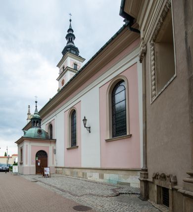 Zdjęcie nr 29: Kościół orientowany, murowany, trzynawowy. Typ halowy, z węższym, prostokątnym prezbiterium zamkniętym trójbocznie oraz z wieżą wbudowaną w zachodnią fasadę. Do prezbiterium od północy przylega zakrystia (do niej od zachodu klatka schodowa wiodąca do skarbca na piętrze), od południa natomiast kaplica Morsztynów z XVII wieku. Kaplica na planie prostokąta zamkniętego trójbocznie, z niszami w ściętych narożach, pod nią sklepiona krypta grobowa, opięta pilastrami wewnątrz i na zewnątrz. Od południa do korpusu przylega kruchta stanowiąca również kaplicę Matki Boskiej Pocieszenia. Korpus czteroprzęsłowy, nawy oddzielają arkady wsparte na filarach, pierwsze stanowią podparcie dla chóru. Nad nawami i prezbiterium strop imitujący sklepienie, na którym malowana dekoracja imitująca gurty, plafony oraz stiuki. Dekoracja ścian powtarza podziały wyznaczone przez arkady, namalowano na nich pilastry podtrzymujące belkowanie. W prezbiterium dwa okna usytuowane niedaleko ołtarza, w nawie osiem, wszystkie w kształcie stojącego prostokąta zamkniętego półkoliście, wypełnione witrażami z dekoracją geometryczną. Główne wejście od zachodu, drugie od północy, trzecie symetrycznie od południa przez kruchtę sklepioną kopułą, kolejne trzy do prezbiterium, od południa przez zakrystię i od północy przez kruchtę. 
Fasada zachodnia na cokole, podzielona gzymsami i lizenami, z wieżą zakończoną hełmem. Na osi drzwi w kształcie stojącego prostokąta flankowane prostokątnymi oknami, powyżej trzecie, większe okno zamknięte łukiem odcinkowym; kondygnację wieńczy gzyms. Druga kondygnacja powtarza podziały pierwszej. W środkowej osi okulus, powyżej dwudzielne okno zamknięte półkoliście. Nad nim tarcza zegara, kolejne dwie na ścianach bocznych. Kondygnacja ograniczona po bokach spływali wolutowymi i obeliskami zakończonymi kulami; przechodzi w wieżę. Powyżej gzyms podtrzymywany kroksztynami. Ostatnia kondygnacja o artykulacji ramowej z balkonami z każdej strony, powyżej wieńczącego gzymsu. Pozostałe elewacje na cokole, kontynuują podziały  lizenowo-ramowe, artykulacją podkreślają okna. Kaplica Morsztynów opięta pilastrami z głowicami, prezbiterium oszkarpowane. Dach nad nawą główną, kaplicą i prezbiterium dwuspadowy, na kruchtą południową kopuła, na wieży ozdobny hełm dołem wklęsły, dekorowany wolutami. Powyżej kopuła, na której latarnia przepruta arkadami i zwieńczona przewężoną w środku kopułą zakończoną ostrosłupem z kulą i krzyżem. 
Do najcenniejszych zabytków świątyni należą m.in. zaprojektowana przez Jana Delamarsa kaplica Morsztynów dekorowana stiukami z 1693 roku autorstwa Baltazara Fontany. Wewnątrz niej warto zwrócić uwagę na osiemnastowieczny ołtarz z przedstawieniem Oblicza Chrystusa i rzeźbionym antepedium. W ołtarz Ukrzyżowania znajdujący się w północnej nawie kościoła wkomponowano gotyckie rzeźby św. Jana i Marii. 