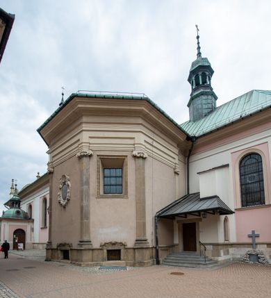 Zdjęcie nr 25: Kościół orientowany, murowany, trzynawowy. Typ halowy, z węższym, prostokątnym prezbiterium zamkniętym trójbocznie oraz z wieżą wbudowaną w zachodnią fasadę. Do prezbiterium od północy przylega zakrystia (do niej od zachodu klatka schodowa wiodąca do skarbca na piętrze), od południa natomiast kaplica Morsztynów z XVII wieku. Kaplica na planie prostokąta zamkniętego trójbocznie, z niszami w ściętych narożach, pod nią sklepiona krypta grobowa, opięta pilastrami wewnątrz i na zewnątrz. Od południa do korpusu przylega kruchta stanowiąca również kaplicę Matki Boskiej Pocieszenia. Korpus czteroprzęsłowy, nawy oddzielają arkady wsparte na filarach, pierwsze stanowią podparcie dla chóru. Nad nawami i prezbiterium strop imitujący sklepienie, na którym malowana dekoracja imitująca gurty, plafony oraz stiuki. Dekoracja ścian powtarza podziały wyznaczone przez arkady, namalowano na nich pilastry podtrzymujące belkowanie. W prezbiterium dwa okna usytuowane niedaleko ołtarza, w nawie osiem, wszystkie w kształcie stojącego prostokąta zamkniętego półkoliście, wypełnione witrażami z dekoracją geometryczną. Główne wejście od zachodu, drugie od północy, trzecie symetrycznie od południa przez kruchtę sklepioną kopułą, kolejne trzy do prezbiterium, od południa przez zakrystię i od północy przez kruchtę. 
Fasada zachodnia na cokole, podzielona gzymsami i lizenami, z wieżą zakończoną hełmem. Na osi drzwi w kształcie stojącego prostokąta flankowane prostokątnymi oknami, powyżej trzecie, większe okno zamknięte łukiem odcinkowym; kondygnację wieńczy gzyms. Druga kondygnacja powtarza podziały pierwszej. W środkowej osi okulus, powyżej dwudzielne okno zamknięte półkoliście. Nad nim tarcza zegara, kolejne dwie na ścianach bocznych. Kondygnacja ograniczona po bokach spływali wolutowymi i obeliskami zakończonymi kulami; przechodzi w wieżę. Powyżej gzyms podtrzymywany kroksztynami. Ostatnia kondygnacja o artykulacji ramowej z balkonami z każdej strony, powyżej wieńczącego gzymsu. Pozostałe elewacje na cokole, kontynuują podziały  lizenowo-ramowe, artykulacją podkreślają okna. Kaplica Morsztynów opięta pilastrami z głowicami, prezbiterium oszkarpowane. Dach nad nawą główną, kaplicą i prezbiterium dwuspadowy, na kruchtą południową kopuła, na wieży ozdobny hełm dołem wklęsły, dekorowany wolutami. Powyżej kopuła, na której latarnia przepruta arkadami i zwieńczona przewężoną w środku kopułą zakończoną ostrosłupem z kulą i krzyżem. 
Do najcenniejszych zabytków świątyni należą m.in. zaprojektowana przez Jana Delamarsa kaplica Morsztynów dekorowana stiukami z 1693 roku autorstwa Baltazara Fontany. Wewnątrz niej warto zwrócić uwagę na osiemnastowieczny ołtarz z przedstawieniem Oblicza Chrystusa i rzeźbionym antepedium. W ołtarz Ukrzyżowania znajdujący się w północnej nawie kościoła wkomponowano gotyckie rzeźby św. Jana i Marii. 