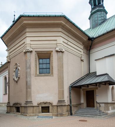 Zdjęcie nr 24: Kościół orientowany, murowany, trzynawowy. Typ halowy, z węższym, prostokątnym prezbiterium zamkniętym trójbocznie oraz z wieżą wbudowaną w zachodnią fasadę. Do prezbiterium od północy przylega zakrystia (do niej od zachodu klatka schodowa wiodąca do skarbca na piętrze), od południa natomiast kaplica Morsztynów z XVII wieku. Kaplica na planie prostokąta zamkniętego trójbocznie, z niszami w ściętych narożach, pod nią sklepiona krypta grobowa, opięta pilastrami wewnątrz i na zewnątrz. Od południa do korpusu przylega kruchta stanowiąca również kaplicę Matki Boskiej Pocieszenia. Korpus czteroprzęsłowy, nawy oddzielają arkady wsparte na filarach, pierwsze stanowią podparcie dla chóru. Nad nawami i prezbiterium strop imitujący sklepienie, na którym malowana dekoracja imitująca gurty, plafony oraz stiuki. Dekoracja ścian powtarza podziały wyznaczone przez arkady, namalowano na nich pilastry podtrzymujące belkowanie. W prezbiterium dwa okna usytuowane niedaleko ołtarza, w nawie osiem, wszystkie w kształcie stojącego prostokąta zamkniętego półkoliście, wypełnione witrażami z dekoracją geometryczną. Główne wejście od zachodu, drugie od północy, trzecie symetrycznie od południa przez kruchtę sklepioną kopułą, kolejne trzy do prezbiterium, od południa przez zakrystię i od północy przez kruchtę. 
Fasada zachodnia na cokole, podzielona gzymsami i lizenami, z wieżą zakończoną hełmem. Na osi drzwi w kształcie stojącego prostokąta flankowane prostokątnymi oknami, powyżej trzecie, większe okno zamknięte łukiem odcinkowym; kondygnację wieńczy gzyms. Druga kondygnacja powtarza podziały pierwszej. W środkowej osi okulus, powyżej dwudzielne okno zamknięte półkoliście. Nad nim tarcza zegara, kolejne dwie na ścianach bocznych. Kondygnacja ograniczona po bokach spływali wolutowymi i obeliskami zakończonymi kulami; przechodzi w wieżę. Powyżej gzyms podtrzymywany kroksztynami. Ostatnia kondygnacja o artykulacji ramowej z balkonami z każdej strony, powyżej wieńczącego gzymsu. Pozostałe elewacje na cokole, kontynuują podziały  lizenowo-ramowe, artykulacją podkreślają okna. Kaplica Morsztynów opięta pilastrami z głowicami, prezbiterium oszkarpowane. Dach nad nawą główną, kaplicą i prezbiterium dwuspadowy, na kruchtą południową kopuła, na wieży ozdobny hełm dołem wklęsły, dekorowany wolutami. Powyżej kopuła, na której latarnia przepruta arkadami i zwieńczona przewężoną w środku kopułą zakończoną ostrosłupem z kulą i krzyżem. 
Do najcenniejszych zabytków świątyni należą m.in. zaprojektowana przez Jana Delamarsa kaplica Morsztynów dekorowana stiukami z 1693 roku autorstwa Baltazara Fontany. Wewnątrz niej warto zwrócić uwagę na osiemnastowieczny ołtarz z przedstawieniem Oblicza Chrystusa i rzeźbionym antepedium. W ołtarz Ukrzyżowania znajdujący się w północnej nawie kościoła wkomponowano gotyckie rzeźby św. Jana i Marii. 