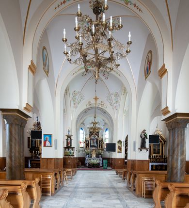 Zdjęcie nr 10: Kościół jest położony w centrum wsi, na północ od główne drogi we wsi, na placu otoczonym kamiennym murem, nakrytym betonowym daszkiem, obsadzony ozdobnym drzewostanem. Wejście na plac kościelny prowadzi przez bramę od południa i wąską furtkę od zachodu. Kościół jest budowlą założoną na planie krzyża łacińskiego, murowaną, skierowaną na północ, trójnawową z transeptem i prezbiterium zakończonym wielobocznie. Wewnątrz dwuprzęsłowe nawy oddzielone są ostrołukowymi arkadami wspartymi na zdwojonych filarach oraz na parze kolumn pośrodku świątyni. Podpory wspierają sklepienie krzyżowo-żebrowe. Od fasady dostawiona masywna wieża na planie prostokąta, zwężająca się ku górze, nakryta namiotowym daszkiem i zwieńczona krzyżem. Wejście południowe w formie prostokątnego portalu, ujętego w dużą ostrołukową arkadę wykonaną z kamiennej okładziny. Powyżej na tle trójdzielnego okna ustawiona jest figura Chrystusa. Pomiędzy zachodnim ramieniem transeptu, a prezbiterium dostawiona zakrystia z wejściem od północy, analogicznie po drugiej stronie kościoła kaplica Najświętszego Serca Pana Jezusa. Nad skrzyżowaniem ramion transeptu umieszczona jest ażurowa wieżyczka na sygnaturkę. Całość opięta szkarpami. Detale dekoracyjne, takie jak narożniki, portale, gzymsy i obramowania okien wykonane z piaskowca. Do świątyni prowadzi też zachodni przedsionek.