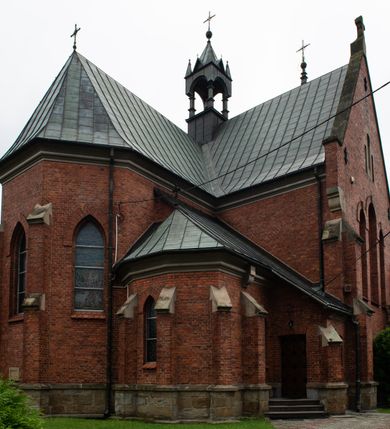 Zdjęcie nr 8: Kościół jest położony w centrum wsi, na północ od główne drogi we wsi, na placu otoczonym kamiennym murem, nakrytym betonowym daszkiem, obsadzony ozdobnym drzewostanem. Wejście na plac kościelny prowadzi przez bramę od południa i wąską furtkę od zachodu. Kościół jest budowlą założoną na planie krzyża łacińskiego, murowaną, skierowaną na północ, trójnawową z transeptem i prezbiterium zakończonym wielobocznie. Wewnątrz dwuprzęsłowe nawy oddzielone są ostrołukowymi arkadami wspartymi na zdwojonych filarach oraz na parze kolumn pośrodku świątyni. Podpory wspierają sklepienie krzyżowo-żebrowe. Od fasady dostawiona masywna wieża na planie prostokąta, zwężająca się ku górze, nakryta namiotowym daszkiem i zwieńczona krzyżem. Wejście południowe w formie prostokątnego portalu, ujętego w dużą ostrołukową arkadę wykonaną z kamiennej okładziny. Powyżej na tle trójdzielnego okna ustawiona jest figura Chrystusa. Pomiędzy zachodnim ramieniem transeptu, a prezbiterium dostawiona zakrystia z wejściem od północy, analogicznie po drugiej stronie kościoła kaplica Najświętszego Serca Pana Jezusa. Nad skrzyżowaniem ramion transeptu umieszczona jest ażurowa wieżyczka na sygnaturkę. Całość opięta szkarpami. Detale dekoracyjne, takie jak narożniki, portale, gzymsy i obramowania okien wykonane z piaskowca. Do świątyni prowadzi też zachodni przedsionek.