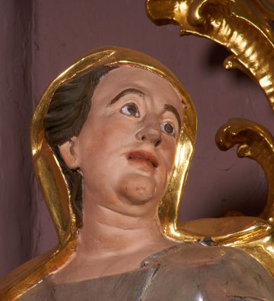 Zdjęcie nr 3: Rzeźba jest drążona z tyłu, ustawiona na niewielkiej, prostej podstawie. Anna przedstawiona w całej postaci, ustawiona frontalnie, z głową skierowaną w lewo, stoi w kontrapoście, lekko odchylona w tył, obie dłonie ma złożone na piersi. Jej twarz posiada rysy dojrzałej kobiety, duże oczy z podniesionymi brwiami, niewielki nos, pełne usta oraz wyodrębnioną brodę z dołeczkiem i drugim podbródkiem. Głowa okolona jest brązowymi włosami ukrytymi częściowo pod płaszczem. Święta ubrana jest w długą tunikę przepasaną w talii, przylegającą do ciała oraz w płaszcz nakrywający głowę, zawiązany z przodu, na jej prawym biodrze. Figura polichromowana jest naturalistycznie w partiach ciała, tunika jest srebrzona, a płaszcz złocony. 