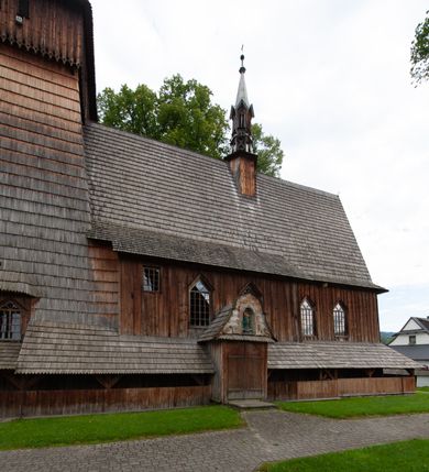 Zdjęcie nr 57: Kościół drewniany, orientowany, składający się z babińca, nawy głównej z przylegającą do niej od północy kaplicą boczną i kruchtą od południa oraz prezbiterium z zakrystią od północy. Prezbiterium i nawa w konstrukcji zrębowej, wieża, soboty i kruchta w słupowej. Kościół jednonawowy, nawa prawie kwadratowa, prezbiterium prostokątne, zamknięte prosto, oddzielone ścianą tęczową. Otwór tęczowy zamknięty łukiem dwuramiennym z biegnącą w poprzek grubą belką. Ściany gładkie, pokryte dekoracją malarską imitującą poziomo ułożone belki z uszczelniającymi je kiczkami, scenami figuralnymi, zacheuszkami i pasami ornamentalnymi. W nawie od zachodu chór muzyczny wsparty na dwóch, profilowanych słupach, z parapetem wybrzuszonym pośrodku; z dekoracją malarską o motywach geometryczno-roślinnych. Nawa i prezbiterium naryte pozornym sklepieniem z pułapami lekko zaokrąglonymi po bokach. Na całości przekrycia dekoracja malarska. W prezbiterium są to postacie aniołów wśród stylizowanych kwiatów na stropie adorujących monogram maryjny, nawę dekorują motywy roślinne. Anioły zdobią również sklepienie kaplicy. Strop babińca oraz fragmenty prezbiterium pokrywa oryginalna dekoracja patronowa złożona z motywów geometrycznych oraz figuralnych. W nawie głównej trzy okna, w prezbiterium i babińcu po dwa – od południa, kolejne do zakrystii; zamknięte łukiem dwuspadowym. Główne wejście od zachodu, drugie przez kruchtę od południa, trzecie do zakrystii. Portal południowy ostrołukowy, dekorowany ornamentem sznurowym, portal do zakrystii zamknięty łukiem w ośli grzbiet. Od zachodu kwadratowa wieża o pochyłych ścianach, z nadwieszoną izbicą, zwieńczona czteropołaciowym, ostrosłupowym hełmem gontowym. Izbica wieży oszalowana dookoła deskami zakończonymi u dołu ozdobnie wyrzynaną koronką. Od południa i wschodu soboty, czyli rodzaj podcieni,  w pozostałych miejscach gontowy kołnierz. Ściany świątyni oraz kaplicy są od zewnątrz szalowane deskami, wieża oraz przedsionek obite są gontem. Dachy i zadaszenia sobot pokryte gontem, kopuła kaplicy oraz wieżyczka na sygnaturkę pokryta blachą. 