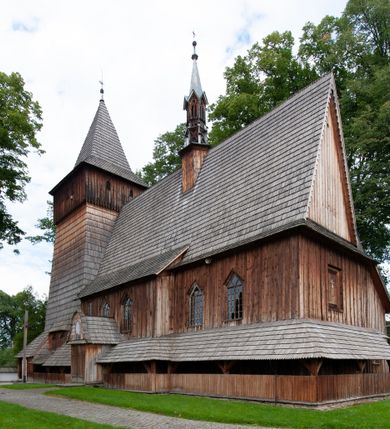 Zdjęcie nr 54: Kościół drewniany, orientowany, składający się z babińca, nawy głównej z przylegającą do niej od północy kaplicą boczną i kruchtą od południa oraz prezbiterium z zakrystią od północy. Prezbiterium i nawa w konstrukcji zrębowej, wieża, soboty i kruchta w słupowej. Kościół jednonawowy, nawa prawie kwadratowa, prezbiterium prostokątne, zamknięte prosto, oddzielone ścianą tęczową. Otwór tęczowy zamknięty łukiem dwuramiennym z biegnącą w poprzek grubą belką. Ściany gładkie, pokryte dekoracją malarską imitującą poziomo ułożone belki z uszczelniającymi je kiczkami, scenami figuralnymi, zacheuszkami i pasami ornamentalnymi. W nawie od zachodu chór muzyczny wsparty na dwóch, profilowanych słupach, z parapetem wybrzuszonym pośrodku; z dekoracją malarską o motywach geometryczno-roślinnych. Nawa i prezbiterium naryte pozornym sklepieniem z pułapami lekko zaokrąglonymi po bokach. Na całości przekrycia dekoracja malarska. W prezbiterium są to postacie aniołów wśród stylizowanych kwiatów na stropie adorujących monogram maryjny, nawę dekorują motywy roślinne. Anioły zdobią również sklepienie kaplicy. Strop babińca oraz fragmenty prezbiterium pokrywa oryginalna dekoracja patronowa złożona z motywów geometrycznych oraz figuralnych. W nawie głównej trzy okna, w prezbiterium i babińcu po dwa – od południa, kolejne do zakrystii; zamknięte łukiem dwuspadowym. Główne wejście od zachodu, drugie przez kruchtę od południa, trzecie do zakrystii. Portal południowy ostrołukowy, dekorowany ornamentem sznurowym, portal do zakrystii zamknięty łukiem w ośli grzbiet. Od zachodu kwadratowa wieża o pochyłych ścianach, z nadwieszoną izbicą, zwieńczona czteropołaciowym, ostrosłupowym hełmem gontowym. Izbica wieży oszalowana dookoła deskami zakończonymi u dołu ozdobnie wyrzynaną koronką. Od południa i wschodu soboty, czyli rodzaj podcieni,  w pozostałych miejscach gontowy kołnierz. Ściany świątyni oraz kaplicy są od zewnątrz szalowane deskami, wieża oraz przedsionek obite są gontem. Dachy i zadaszenia sobot pokryte gontem, kopuła kaplicy oraz wieżyczka na sygnaturkę pokryta blachą. 