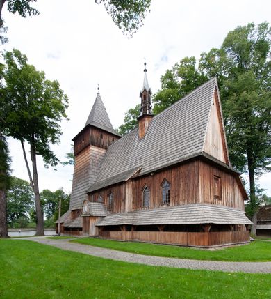 Zdjęcie nr 48: Kościół drewniany, orientowany, składający się z babińca, nawy głównej z przylegającą do niej od północy kaplicą boczną i kruchtą od południa oraz prezbiterium z zakrystią od północy. Prezbiterium i nawa w konstrukcji zrębowej, wieża, soboty i kruchta w słupowej. Kościół jednonawowy, nawa prawie kwadratowa, prezbiterium prostokątne, zamknięte prosto, oddzielone ścianą tęczową. Otwór tęczowy zamknięty łukiem dwuramiennym z biegnącą w poprzek grubą belką. Ściany gładkie, pokryte dekoracją malarską imitującą poziomo ułożone belki z uszczelniającymi je kiczkami, scenami figuralnymi, zacheuszkami i pasami ornamentalnymi. W nawie od zachodu chór muzyczny wsparty na dwóch, profilowanych słupach, z parapetem wybrzuszonym pośrodku; z dekoracją malarską o motywach geometryczno-roślinnych. Nawa i prezbiterium naryte pozornym sklepieniem z pułapami lekko zaokrąglonymi po bokach. Na całości przekrycia dekoracja malarska. W prezbiterium są to postacie aniołów wśród stylizowanych kwiatów na stropie adorujących monogram maryjny, nawę dekorują motywy roślinne. Anioły zdobią również sklepienie kaplicy. Strop babińca oraz fragmenty prezbiterium pokrywa oryginalna dekoracja patronowa złożona z motywów geometrycznych oraz figuralnych. W nawie głównej trzy okna, w prezbiterium i babińcu po dwa – od południa, kolejne do zakrystii; zamknięte łukiem dwuspadowym. Główne wejście od zachodu, drugie przez kruchtę od południa, trzecie do zakrystii. Portal południowy ostrołukowy, dekorowany ornamentem sznurowym, portal do zakrystii zamknięty łukiem w ośli grzbiet. Od zachodu kwadratowa wieża o pochyłych ścianach, z nadwieszoną izbicą, zwieńczona czteropołaciowym, ostrosłupowym hełmem gontowym. Izbica wieży oszalowana dookoła deskami zakończonymi u dołu ozdobnie wyrzynaną koronką. Od południa i wschodu soboty, czyli rodzaj podcieni,  w pozostałych miejscach gontowy kołnierz. Ściany świątyni oraz kaplicy są od zewnątrz szalowane deskami, wieża oraz przedsionek obite są gontem. Dachy i zadaszenia sobot pokryte gontem, kopuła kaplicy oraz wieżyczka na sygnaturkę pokryta blachą. 