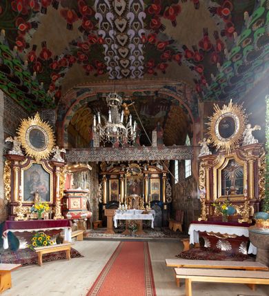 Zdjęcie nr 9: Kościół drewniany, orientowany, składający się z babińca, nawy głównej z przylegającą do niej od północy kaplicą boczną i kruchtą od południa oraz prezbiterium z zakrystią od północy. Prezbiterium i nawa w konstrukcji zrębowej, wieża, soboty i kruchta w słupowej. Kościół jednonawowy, nawa prawie kwadratowa, prezbiterium prostokątne, zamknięte prosto, oddzielone ścianą tęczową. Otwór tęczowy zamknięty łukiem dwuramiennym z biegnącą w poprzek grubą belką. Ściany gładkie, pokryte dekoracją malarską imitującą poziomo ułożone belki z uszczelniającymi je kiczkami, scenami figuralnymi, zacheuszkami i pasami ornamentalnymi. W nawie od zachodu chór muzyczny wsparty na dwóch, profilowanych słupach, z parapetem wybrzuszonym pośrodku; z dekoracją malarską o motywach geometryczno-roślinnych. Nawa i prezbiterium naryte pozornym sklepieniem z pułapami lekko zaokrąglonymi po bokach. Na całości przekrycia dekoracja malarska. W prezbiterium są to postacie aniołów wśród stylizowanych kwiatów na stropie adorujących monogram maryjny, nawę dekorują motywy roślinne. Anioły zdobią również sklepienie kaplicy. Strop babińca oraz fragmenty prezbiterium pokrywa oryginalna dekoracja patronowa złożona z motywów geometrycznych oraz figuralnych. W nawie głównej trzy okna, w prezbiterium i babińcu po dwa – od południa, kolejne do zakrystii; zamknięte łukiem dwuspadowym. Główne wejście od zachodu, drugie przez kruchtę od południa, trzecie do zakrystii. Portal południowy ostrołukowy, dekorowany ornamentem sznurowym, portal do zakrystii zamknięty łukiem w ośli grzbiet. Od zachodu kwadratowa wieża o pochyłych ścianach, z nadwieszoną izbicą, zwieńczona czteropołaciowym, ostrosłupowym hełmem gontowym. Izbica wieży oszalowana dookoła deskami zakończonymi u dołu ozdobnie wyrzynaną koronką. Od południa i wschodu soboty, czyli rodzaj podcieni,  w pozostałych miejscach gontowy kołnierz. Ściany świątyni oraz kaplicy są od zewnątrz szalowane deskami, wieża oraz przedsionek obite są gontem. Dachy i zadaszenia sobot pokryte gontem, kopuła kaplicy oraz wieżyczka na sygnaturkę pokryta blachą. 