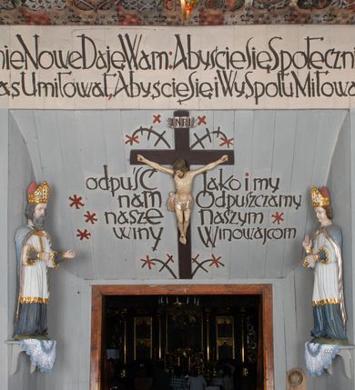 Zdjęcie nr 25: Kościół drewniany, orientowany, składający się z babińca, nawy głównej z przylegającą do niej od północy kaplicą boczną i kruchtą od południa oraz prezbiterium z zakrystią od północy. Prezbiterium i nawa w konstrukcji zrębowej, wieża, soboty i kruchta w słupowej. Kościół jednonawowy, nawa prawie kwadratowa, prezbiterium prostokątne, zamknięte prosto, oddzielone ścianą tęczową. Otwór tęczowy zamknięty łukiem dwuramiennym z biegnącą w poprzek grubą belką. Ściany gładkie, pokryte dekoracją malarską imitującą poziomo ułożone belki z uszczelniającymi je kiczkami, scenami figuralnymi, zacheuszkami i pasami ornamentalnymi. W nawie od zachodu chór muzyczny wsparty na dwóch, profilowanych słupach, z parapetem wybrzuszonym pośrodku; z dekoracją malarską o motywach geometryczno-roślinnych. Nawa i prezbiterium naryte pozornym sklepieniem z pułapami lekko zaokrąglonymi po bokach. Na całości przekrycia dekoracja malarska. W prezbiterium są to postacie aniołów wśród stylizowanych kwiatów na stropie adorujących monogram maryjny, nawę dekorują motywy roślinne. Anioły zdobią również sklepienie kaplicy. Strop babińca oraz fragmenty prezbiterium pokrywa oryginalna dekoracja patronowa złożona z motywów geometrycznych oraz figuralnych. W nawie głównej trzy okna, w prezbiterium i babińcu po dwa – od południa, kolejne do zakrystii; zamknięte łukiem dwuspadowym. Główne wejście od zachodu, drugie przez kruchtę od południa, trzecie do zakrystii. Portal południowy ostrołukowy, dekorowany ornamentem sznurowym, portal do zakrystii zamknięty łukiem w ośli grzbiet. Od zachodu kwadratowa wieża o pochyłych ścianach, z nadwieszoną izbicą, zwieńczona czteropołaciowym, ostrosłupowym hełmem gontowym. Izbica wieży oszalowana dookoła deskami zakończonymi u dołu ozdobnie wyrzynaną koronką. Od południa i wschodu soboty, czyli rodzaj podcieni,  w pozostałych miejscach gontowy kołnierz. Ściany świątyni oraz kaplicy są od zewnątrz szalowane deskami, wieża oraz przedsionek obite są gontem. Dachy i zadaszenia sobot pokryte gontem, kopuła kaplicy oraz wieżyczka na sygnaturkę pokryta blachą. 