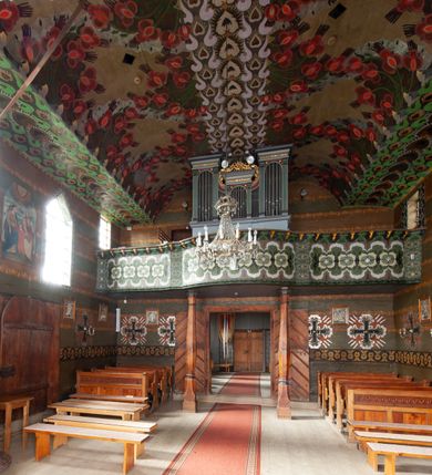 Zdjęcie nr 20: Kościół drewniany, orientowany, składający się z babińca, nawy głównej z przylegającą do niej od północy kaplicą boczną i kruchtą od południa oraz prezbiterium z zakrystią od północy. Prezbiterium i nawa w konstrukcji zrębowej, wieża, soboty i kruchta w słupowej. Kościół jednonawowy, nawa prawie kwadratowa, prezbiterium prostokątne, zamknięte prosto, oddzielone ścianą tęczową. Otwór tęczowy zamknięty łukiem dwuramiennym z biegnącą w poprzek grubą belką. Ściany gładkie, pokryte dekoracją malarską imitującą poziomo ułożone belki z uszczelniającymi je kiczkami, scenami figuralnymi, zacheuszkami i pasami ornamentalnymi. W nawie od zachodu chór muzyczny wsparty na dwóch, profilowanych słupach, z parapetem wybrzuszonym pośrodku; z dekoracją malarską o motywach geometryczno-roślinnych. Nawa i prezbiterium naryte pozornym sklepieniem z pułapami lekko zaokrąglonymi po bokach. Na całości przekrycia dekoracja malarska. W prezbiterium są to postacie aniołów wśród stylizowanych kwiatów na stropie adorujących monogram maryjny, nawę dekorują motywy roślinne. Anioły zdobią również sklepienie kaplicy. Strop babińca oraz fragmenty prezbiterium pokrywa oryginalna dekoracja patronowa złożona z motywów geometrycznych oraz figuralnych. W nawie głównej trzy okna, w prezbiterium i babińcu po dwa – od południa, kolejne do zakrystii; zamknięte łukiem dwuspadowym. Główne wejście od zachodu, drugie przez kruchtę od południa, trzecie do zakrystii. Portal południowy ostrołukowy, dekorowany ornamentem sznurowym, portal do zakrystii zamknięty łukiem w ośli grzbiet. Od zachodu kwadratowa wieża o pochyłych ścianach, z nadwieszoną izbicą, zwieńczona czteropołaciowym, ostrosłupowym hełmem gontowym. Izbica wieży oszalowana dookoła deskami zakończonymi u dołu ozdobnie wyrzynaną koronką. Od południa i wschodu soboty, czyli rodzaj podcieni,  w pozostałych miejscach gontowy kołnierz. Ściany świątyni oraz kaplicy są od zewnątrz szalowane deskami, wieża oraz przedsionek obite są gontem. Dachy i zadaszenia sobot pokryte gontem, kopuła kaplicy oraz wieżyczka na sygnaturkę pokryta blachą. 