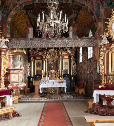 Zdjęcie nr 10: Kościół drewniany, orientowany, składający się z babińca, nawy głównej z przylegającą do niej od północy kaplicą boczną i kruchtą od południa oraz prezbiterium z zakrystią od północy. Prezbiterium i nawa w konstrukcji zrębowej, wieża, soboty i kruchta w słupowej. Kościół jednonawowy, nawa prawie kwadratowa, prezbiterium prostokątne, zamknięte prosto, oddzielone ścianą tęczową. Otwór tęczowy zamknięty łukiem dwuramiennym z biegnącą w poprzek grubą belką. Ściany gładkie, pokryte dekoracją malarską imitującą poziomo ułożone belki z uszczelniającymi je kiczkami, scenami figuralnymi, zacheuszkami i pasami ornamentalnymi. W nawie od zachodu chór muzyczny wsparty na dwóch, profilowanych słupach, z parapetem wybrzuszonym pośrodku; z dekoracją malarską o motywach geometryczno-roślinnych. Nawa i prezbiterium naryte pozornym sklepieniem z pułapami lekko zaokrąglonymi po bokach. Na całości przekrycia dekoracja malarska. W prezbiterium są to postacie aniołów wśród stylizowanych kwiatów na stropie adorujących monogram maryjny, nawę dekorują motywy roślinne. Anioły zdobią również sklepienie kaplicy. Strop babińca oraz fragmenty prezbiterium pokrywa oryginalna dekoracja patronowa złożona z motywów geometrycznych oraz figuralnych. W nawie głównej trzy okna, w prezbiterium i babińcu po dwa – od południa, kolejne do zakrystii; zamknięte łukiem dwuspadowym. Główne wejście od zachodu, drugie przez kruchtę od południa, trzecie do zakrystii. Portal południowy ostrołukowy, dekorowany ornamentem sznurowym, portal do zakrystii zamknięty łukiem w ośli grzbiet. Od zachodu kwadratowa wieża o pochyłych ścianach, z nadwieszoną izbicą, zwieńczona czteropołaciowym, ostrosłupowym hełmem gontowym. Izbica wieży oszalowana dookoła deskami zakończonymi u dołu ozdobnie wyrzynaną koronką. Od południa i wschodu soboty, czyli rodzaj podcieni,  w pozostałych miejscach gontowy kołnierz. Ściany świątyni oraz kaplicy są od zewnątrz szalowane deskami, wieża oraz przedsionek obite są gontem. Dachy i zadaszenia sobot pokryte gontem, kopuła kaplicy oraz wieżyczka na sygnaturkę pokryta blachą. 