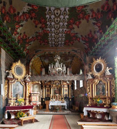 Zdjęcie nr 7: Kościół drewniany, orientowany, składający się z babińca, nawy głównej z przylegającą do niej od północy kaplicą boczną i kruchtą od południa oraz prezbiterium z zakrystią od północy. Prezbiterium i nawa w konstrukcji zrębowej, wieża, soboty i kruchta w słupowej. Kościół jednonawowy, nawa prawie kwadratowa, prezbiterium prostokątne, zamknięte prosto, oddzielone ścianą tęczową. Otwór tęczowy zamknięty łukiem dwuramiennym z biegnącą w poprzek grubą belką. Ściany gładkie, pokryte dekoracją malarską imitującą poziomo ułożone belki z uszczelniającymi je kiczkami, scenami figuralnymi, zacheuszkami i pasami ornamentalnymi. W nawie od zachodu chór muzyczny wsparty na dwóch, profilowanych słupach, z parapetem wybrzuszonym pośrodku; z dekoracją malarską o motywach geometryczno-roślinnych. Nawa i prezbiterium naryte pozornym sklepieniem z pułapami lekko zaokrąglonymi po bokach. Na całości przekrycia dekoracja malarska. W prezbiterium są to postacie aniołów wśród stylizowanych kwiatów na stropie adorujących monogram maryjny, nawę dekorują motywy roślinne. Anioły zdobią również sklepienie kaplicy. Strop babińca oraz fragmenty prezbiterium pokrywa oryginalna dekoracja patronowa złożona z motywów geometrycznych oraz figuralnych. W nawie głównej trzy okna, w prezbiterium i babińcu po dwa – od południa, kolejne do zakrystii; zamknięte łukiem dwuspadowym. Główne wejście od zachodu, drugie przez kruchtę od południa, trzecie do zakrystii. Portal południowy ostrołukowy, dekorowany ornamentem sznurowym, portal do zakrystii zamknięty łukiem w ośli grzbiet. Od zachodu kwadratowa wieża o pochyłych ścianach, z nadwieszoną izbicą, zwieńczona czteropołaciowym, ostrosłupowym hełmem gontowym. Izbica wieży oszalowana dookoła deskami zakończonymi u dołu ozdobnie wyrzynaną koronką. Od południa i wschodu soboty, czyli rodzaj podcieni,  w pozostałych miejscach gontowy kołnierz. Ściany świątyni oraz kaplicy są od zewnątrz szalowane deskami, wieża oraz przedsionek obite są gontem. Dachy i zadaszenia sobot pokryte gontem, kopuła kaplicy oraz wieżyczka na sygnaturkę pokryta blachą. 